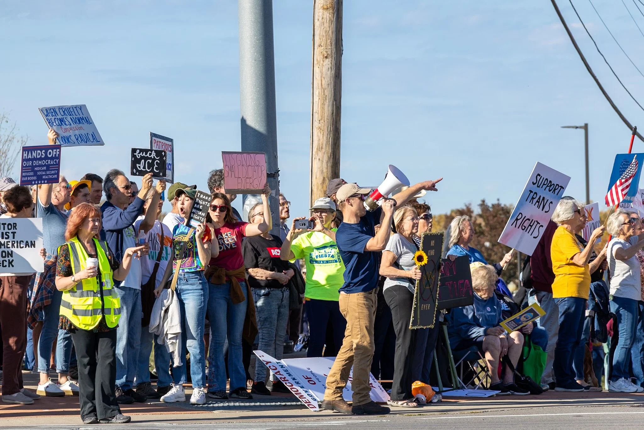 A group of protesters holding various signs and banners during a rally, with some individuals using a megaphone and cameras, outdoors on a sunny day.