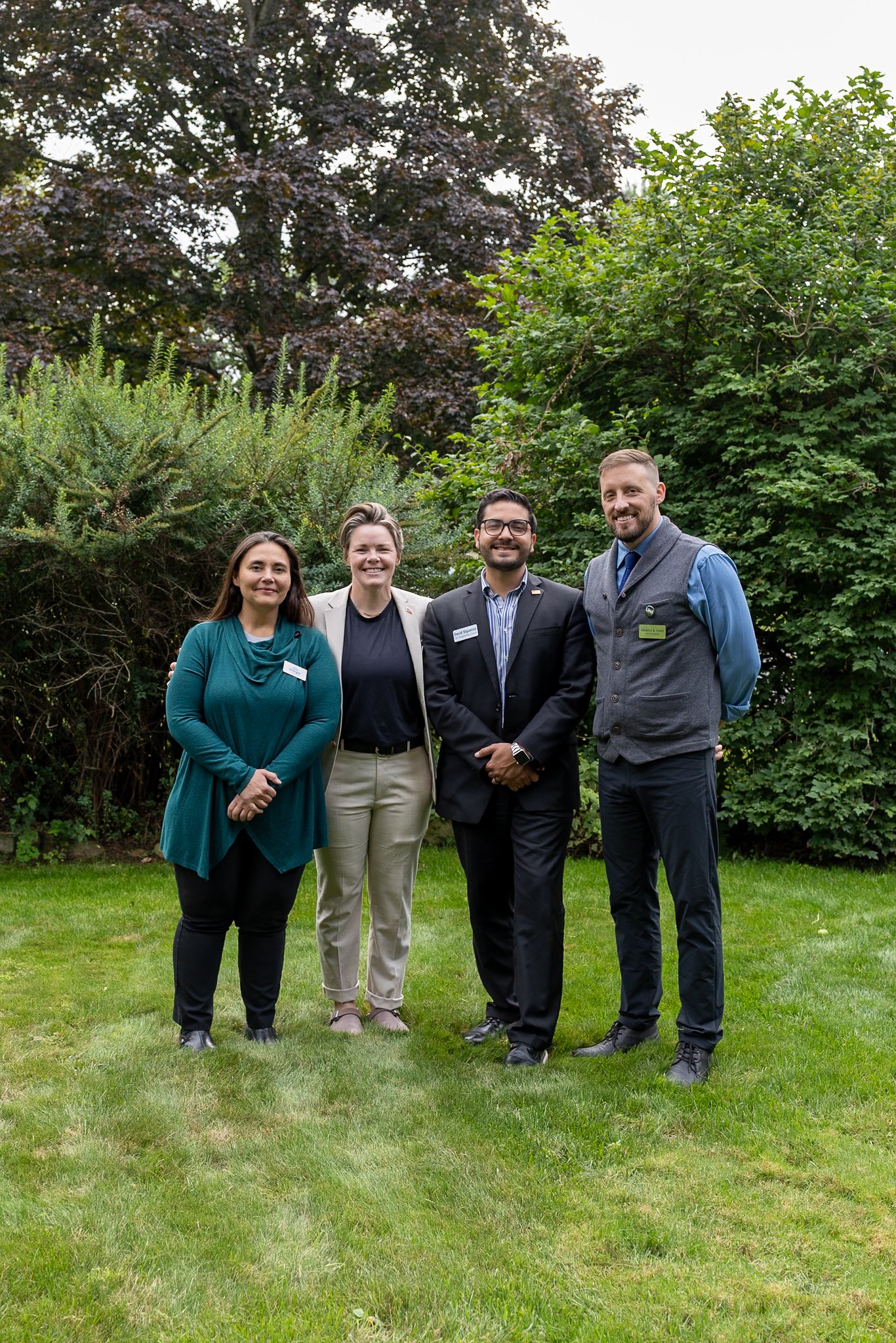 Four people dressed in business attire standing on grass in front of greenery and trees.