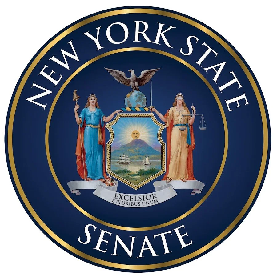 Seal of the New York State Senate featuring two women holding a shield with a mountain, ship, and sun, an eagle on top, and a banner with the Latin motto.