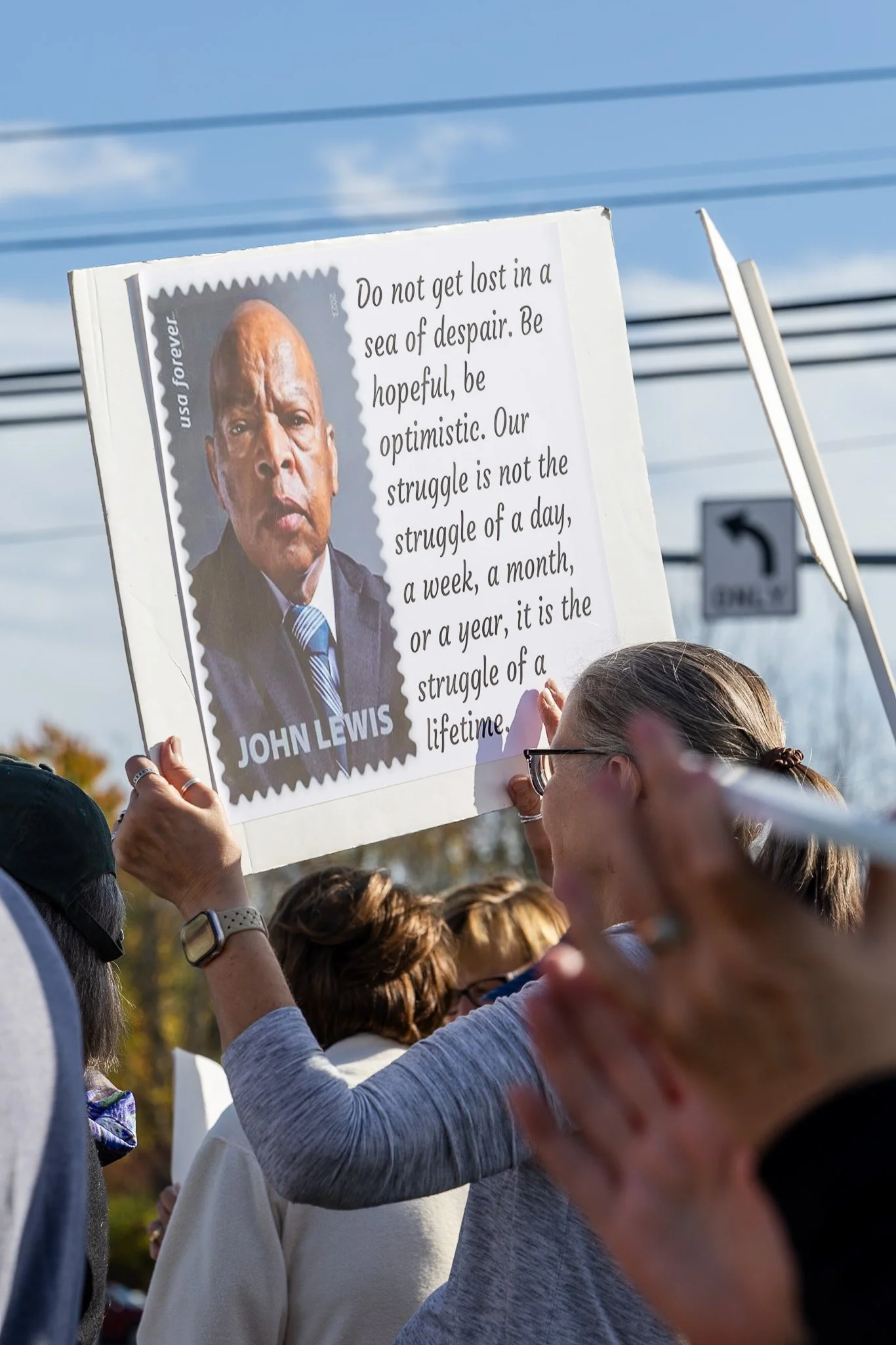 A group of people at a rally holding a sign featuring a photo of John Lewis and a quote about hope and struggle, with power lines and a street sign in the background.