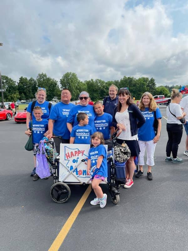 Group of people, including children and adults, standing on a parking lot during a parade or outdoor event. They are holding a sign that reads "HAPPY 4th OF JULY" and some are wearing matching blue shirts with election-related text.