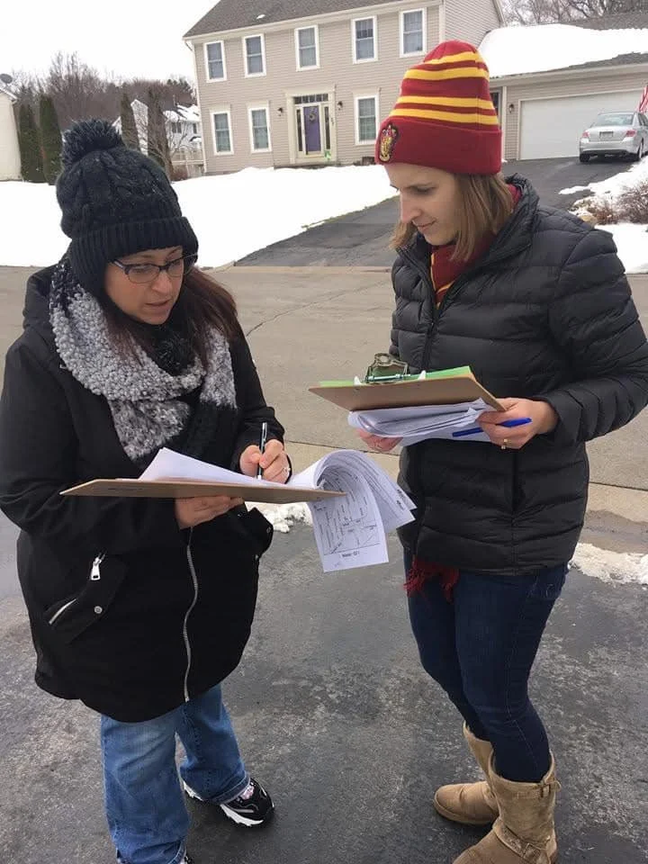 Two women in winter clothing standing on a residential street, looking at papers clipped on clipboards.