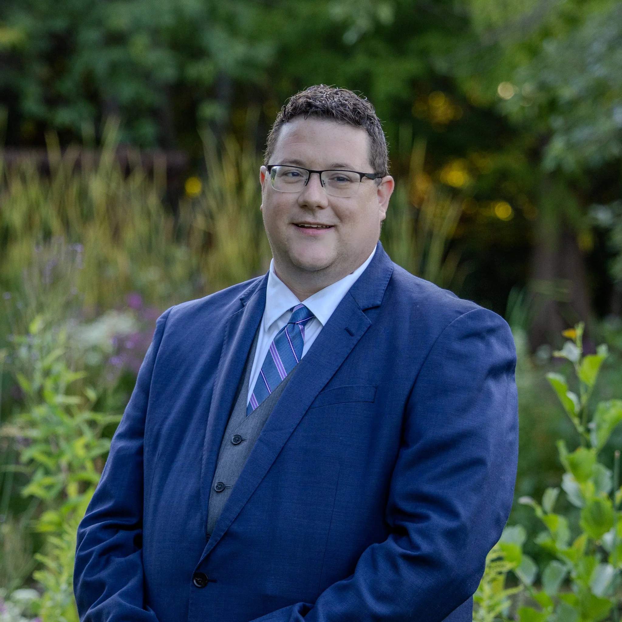 A man dressed in a blue suit with a white shirt and a purple striped tie, standing outdoors in a garden with green foliage and flowering plants in the background, smiling at the camera.