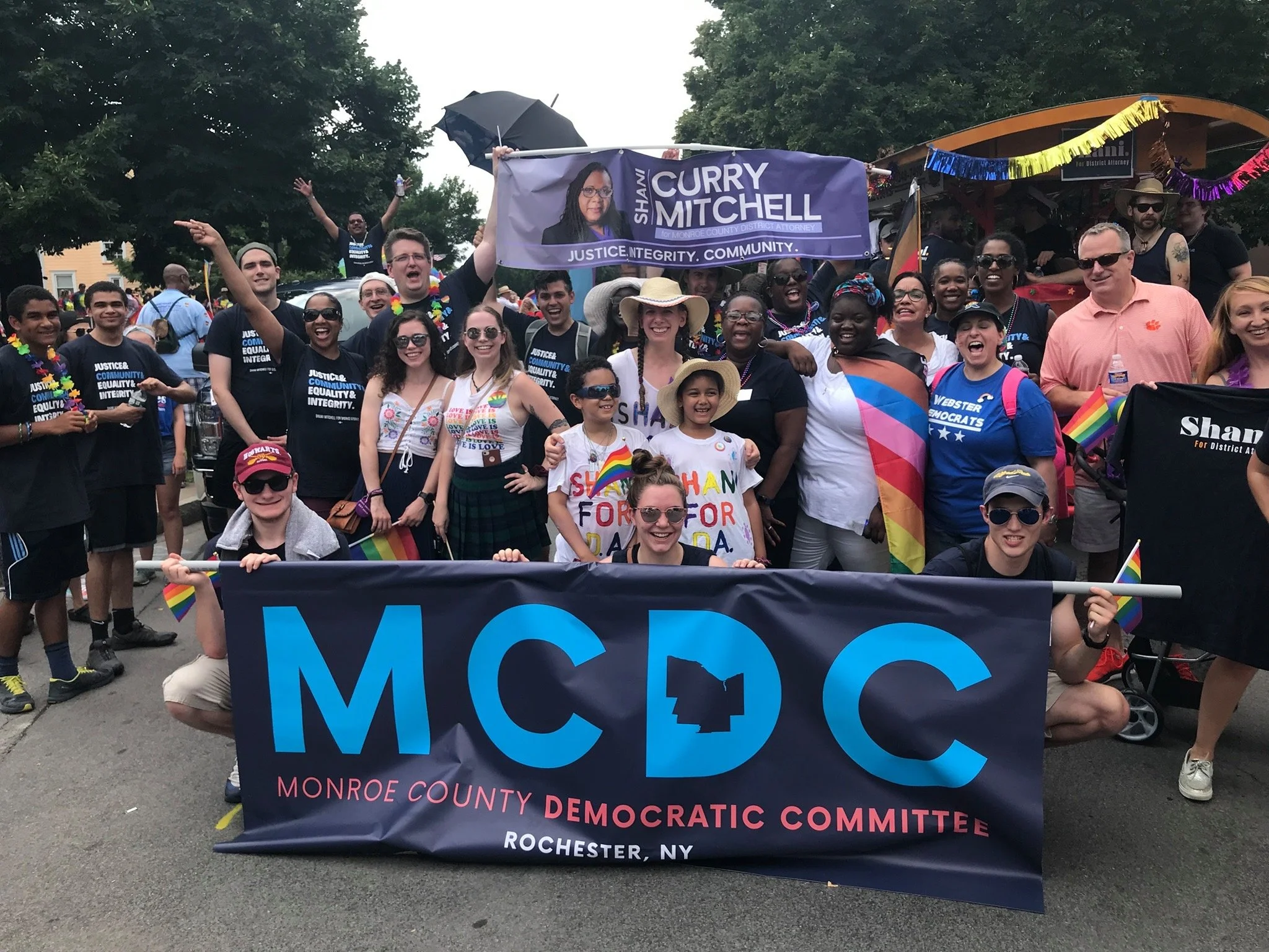 A diverse group of people participating in a pride parade, holding a banner for the Monroe County Democratic Committee, with rainbow flags, colorful clothing, and celebratory expressions.