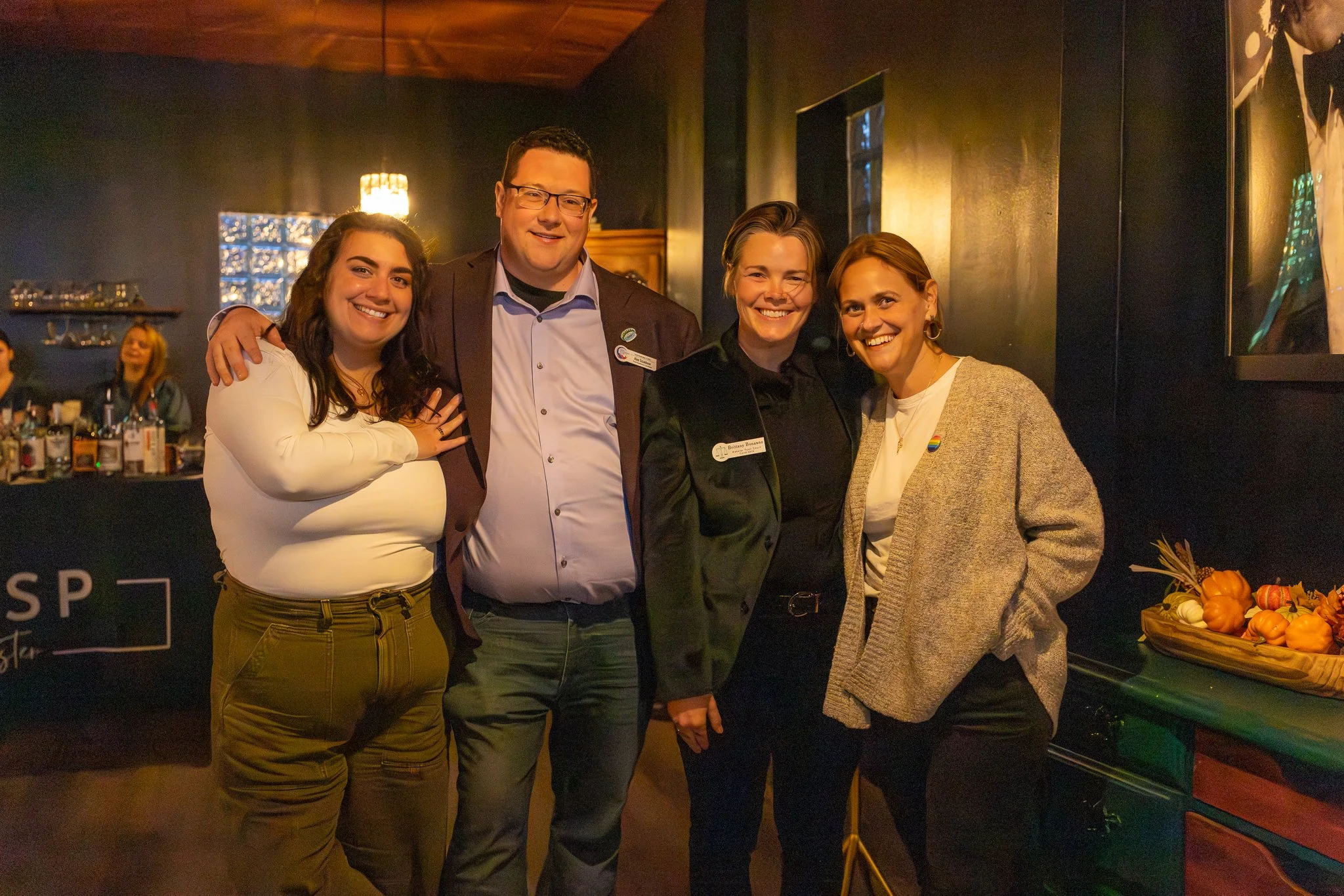 Four people smiling and standing close together indoors, with a bar in the background and a table with pumpkins on the right.
