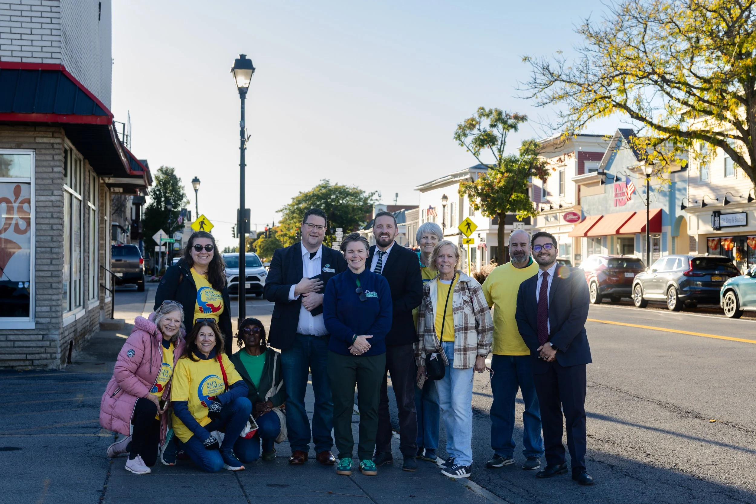 Group of people standing on a sidewalk in a small town, smiling at the camera, some wearing yellow shirts with a logo, with buildings and parked cars in the background.