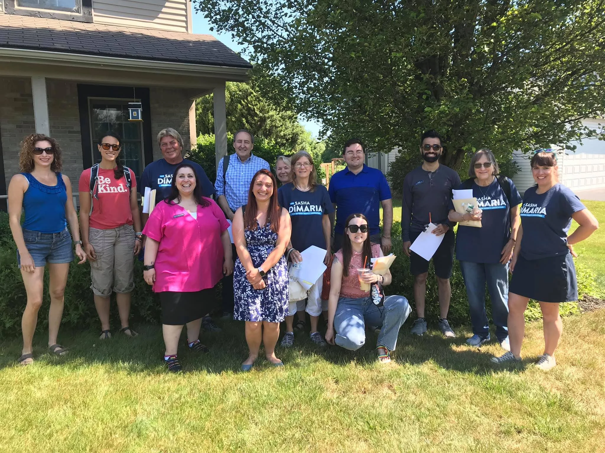Group of 13 people standing outside in front of a house on a sunny day, with some holding papers or drinks, smiling at the camera.