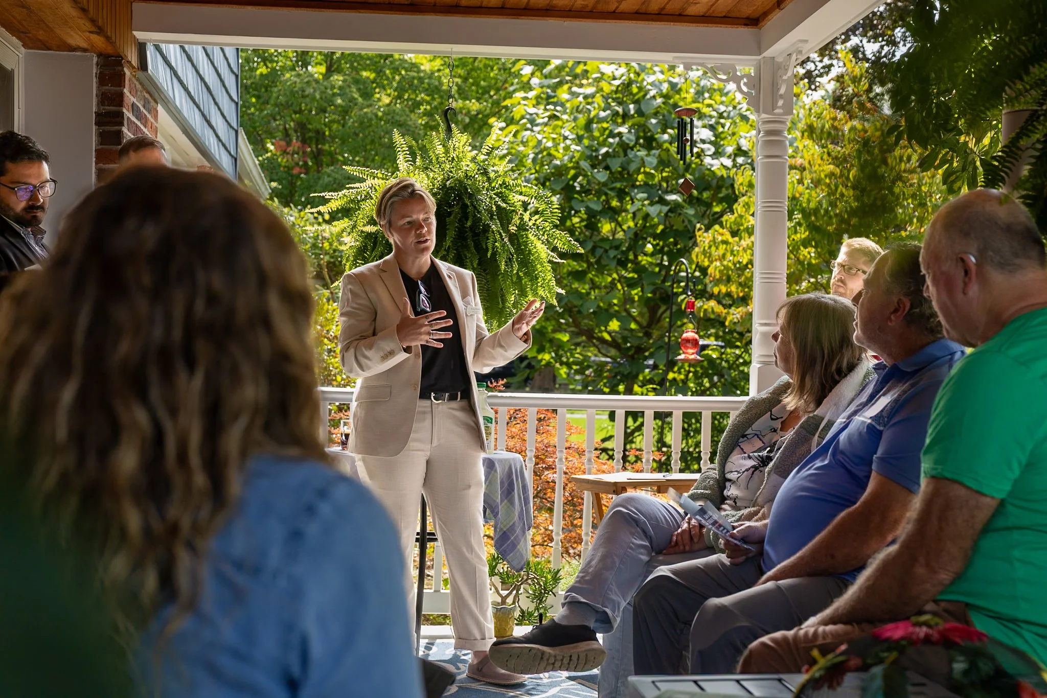 A woman in a beige suit giving a presentation to a group of people on a porch surrounded by greenery.