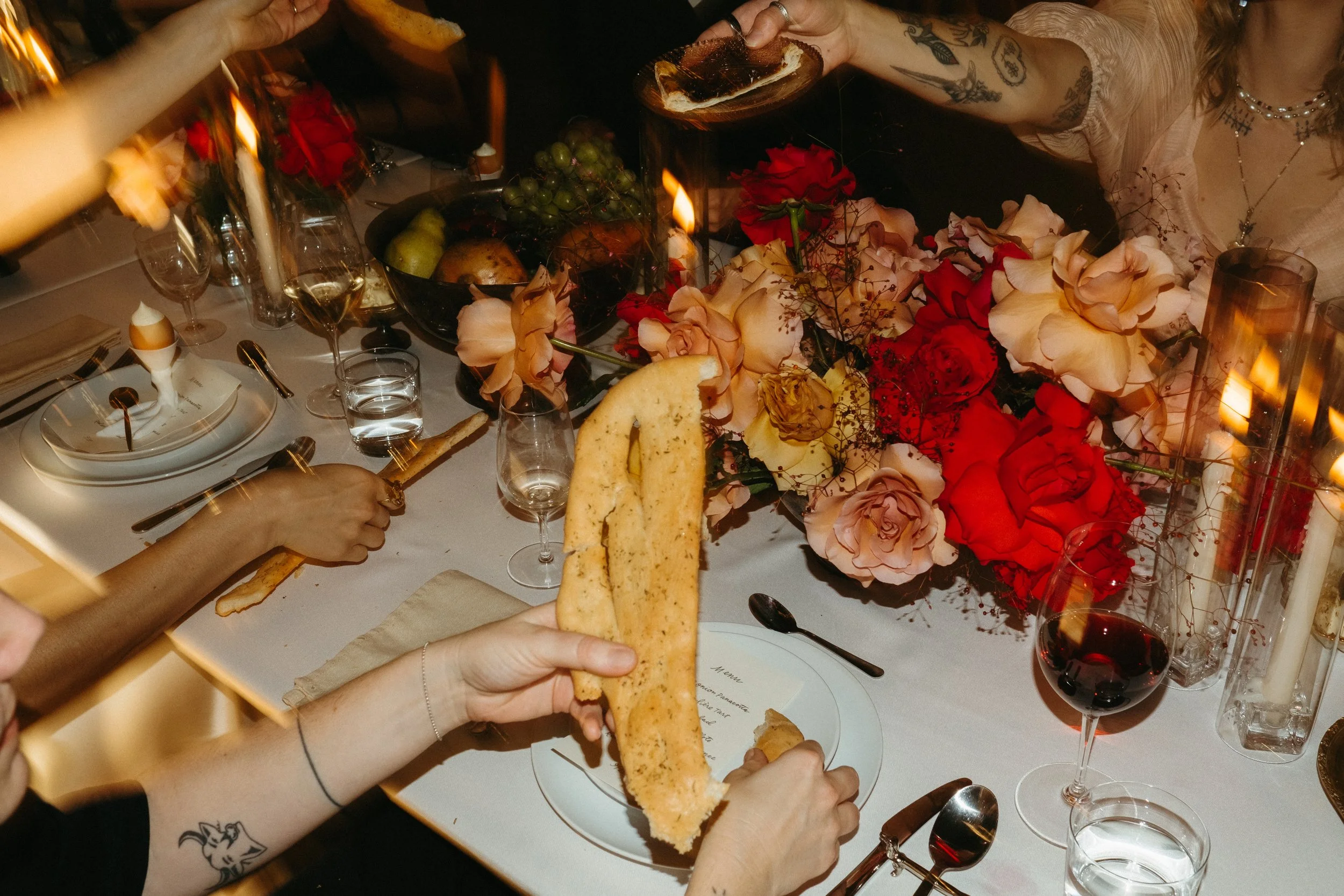People set a table for a celebration with a large bouquet of red and blush roses, glasses of wine, and a person holding a slice of bread.