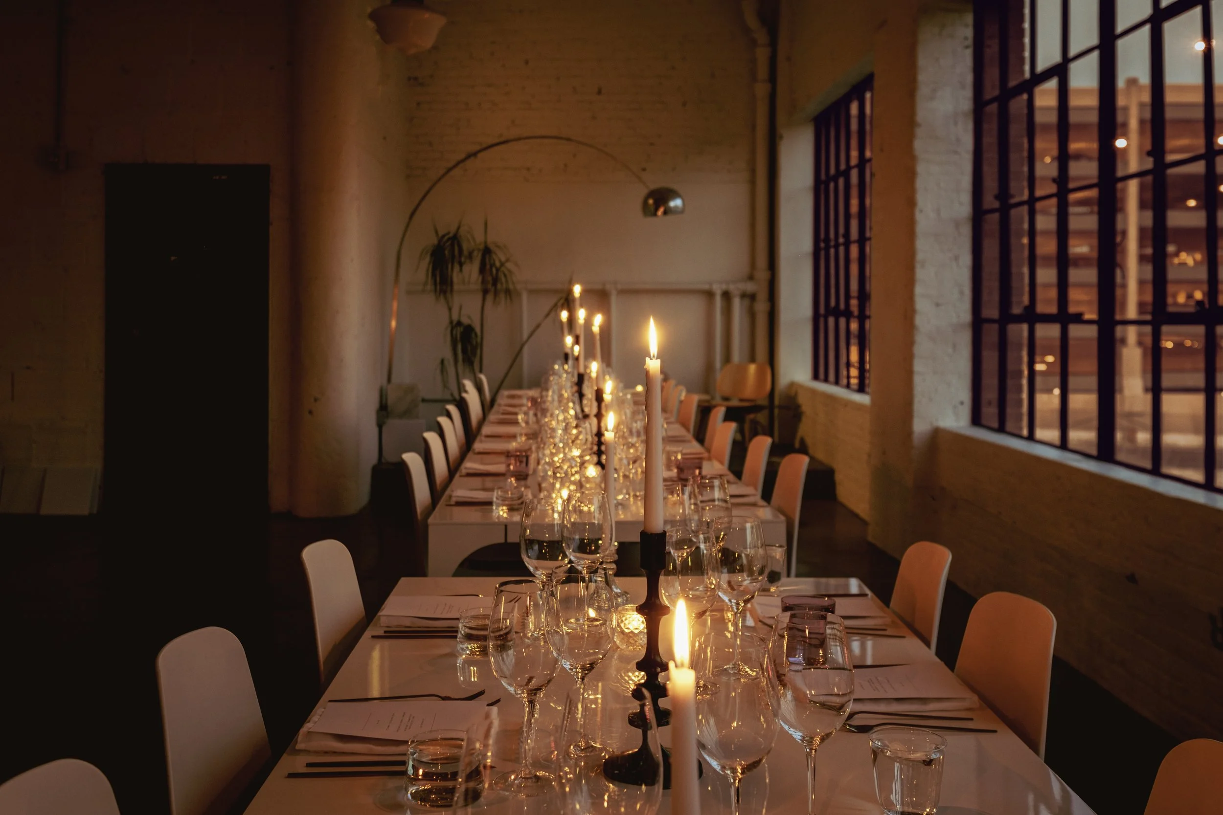 A long dining table decorated with candles, glassware, plates, and utensils inside a dimly lit restaurant with large windows and exposed brick walls.