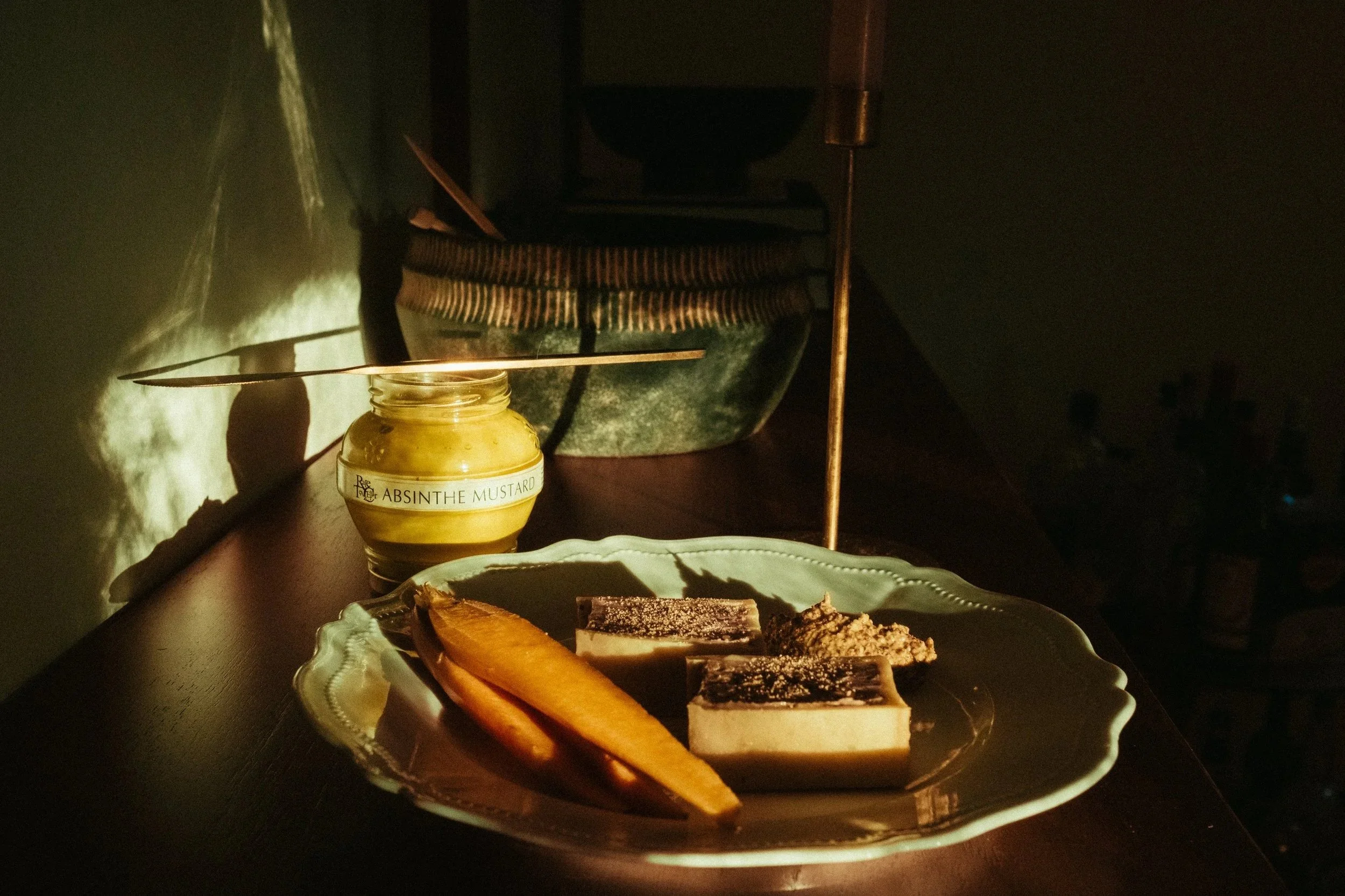 A plate with dessert and slices of dried fruit on a dark wooden table, with a jar of mustard labeled 'Absinthe Mustard' in the background; dim lighting and cozy ambiance.