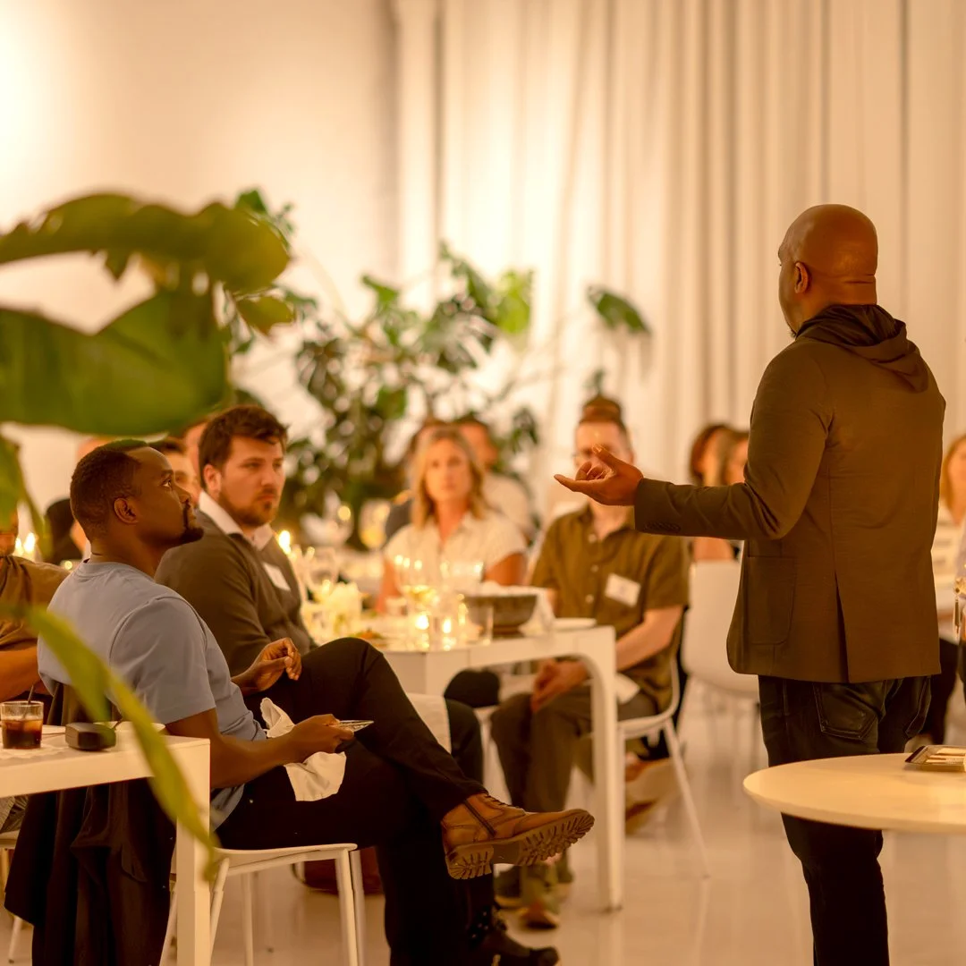 A man is giving a presentation to a group of people seated around a dining table in a well-lit room decorated with plants.