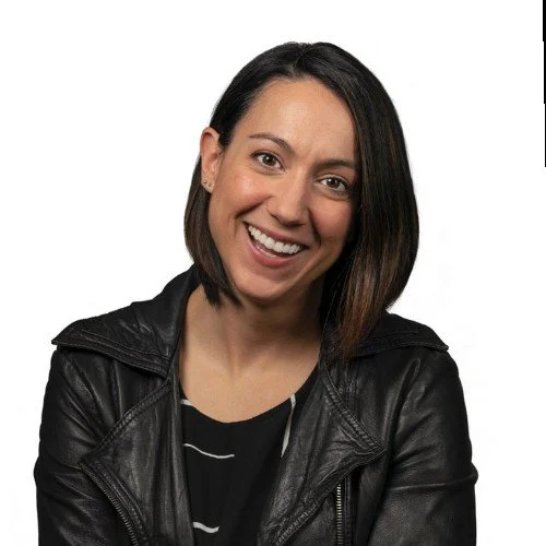 Woman with shoulder-length brown hair smiling, wearing a black leather jacket and a black and white striped shirt, against a white background.