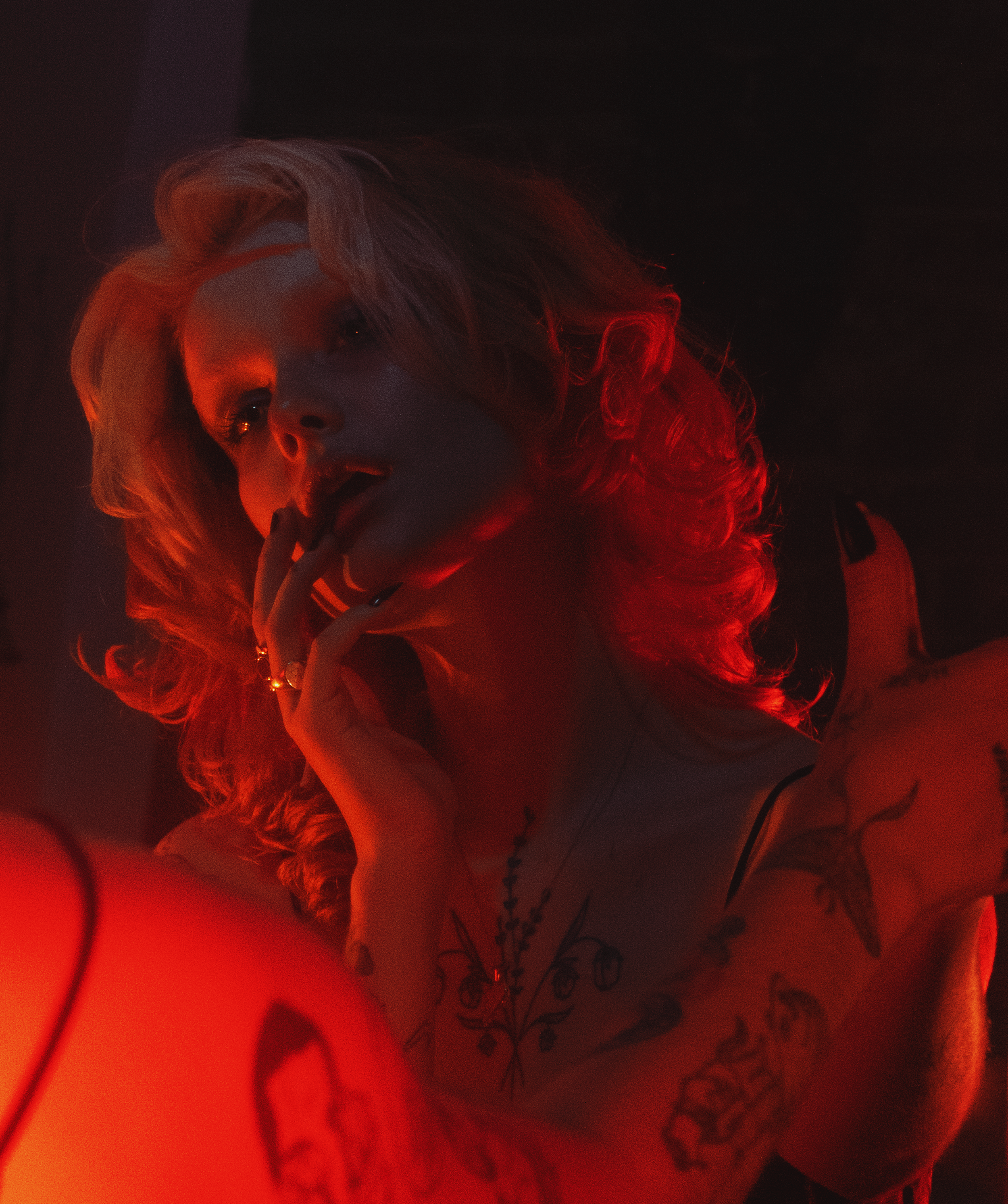 A woman with long curly hair, illuminated in red light, holding a cigarette near her mouth.