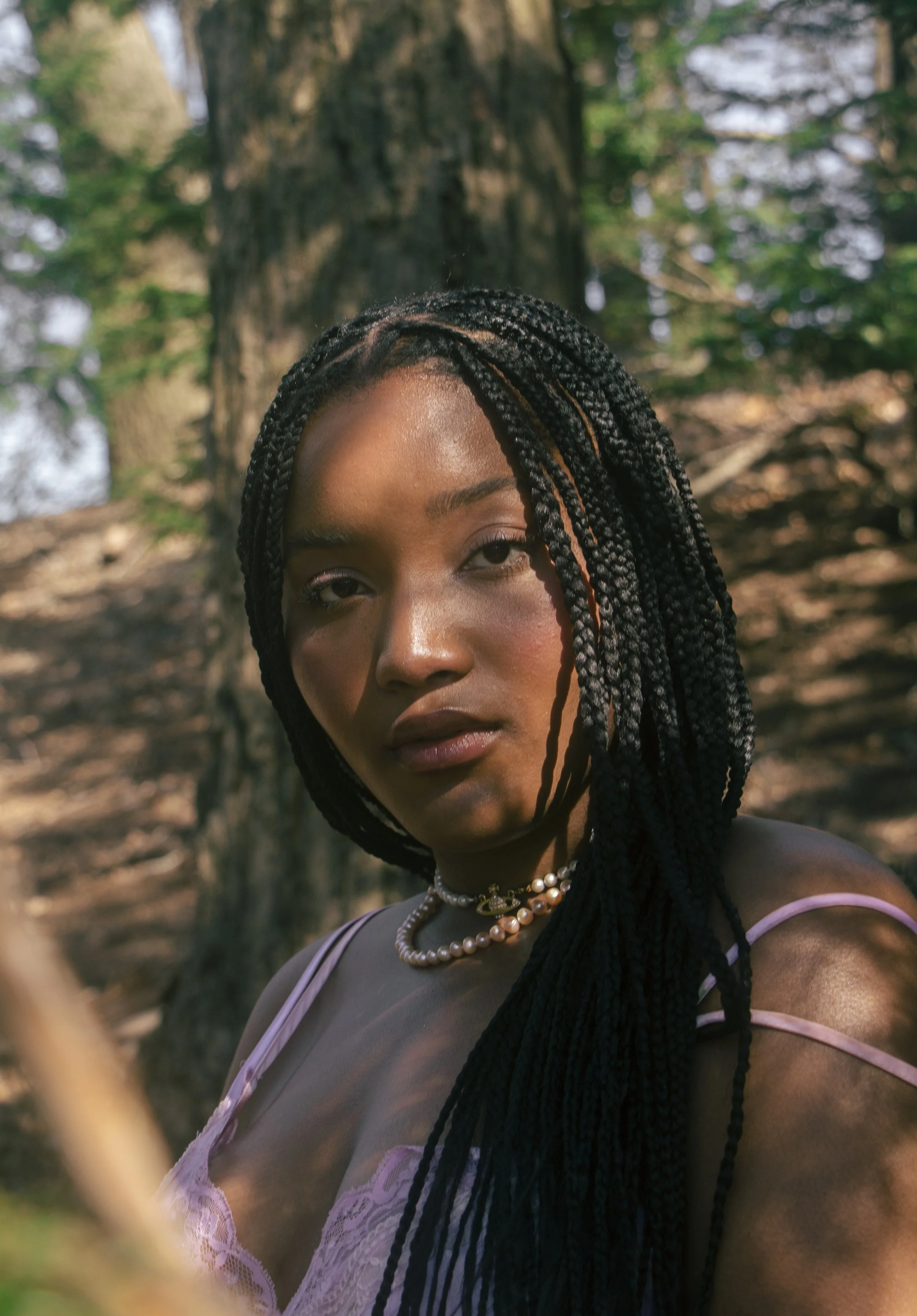 A young woman with braided black hair outdoors in a forest, wearing a pink lace dress and pearl jewelry.