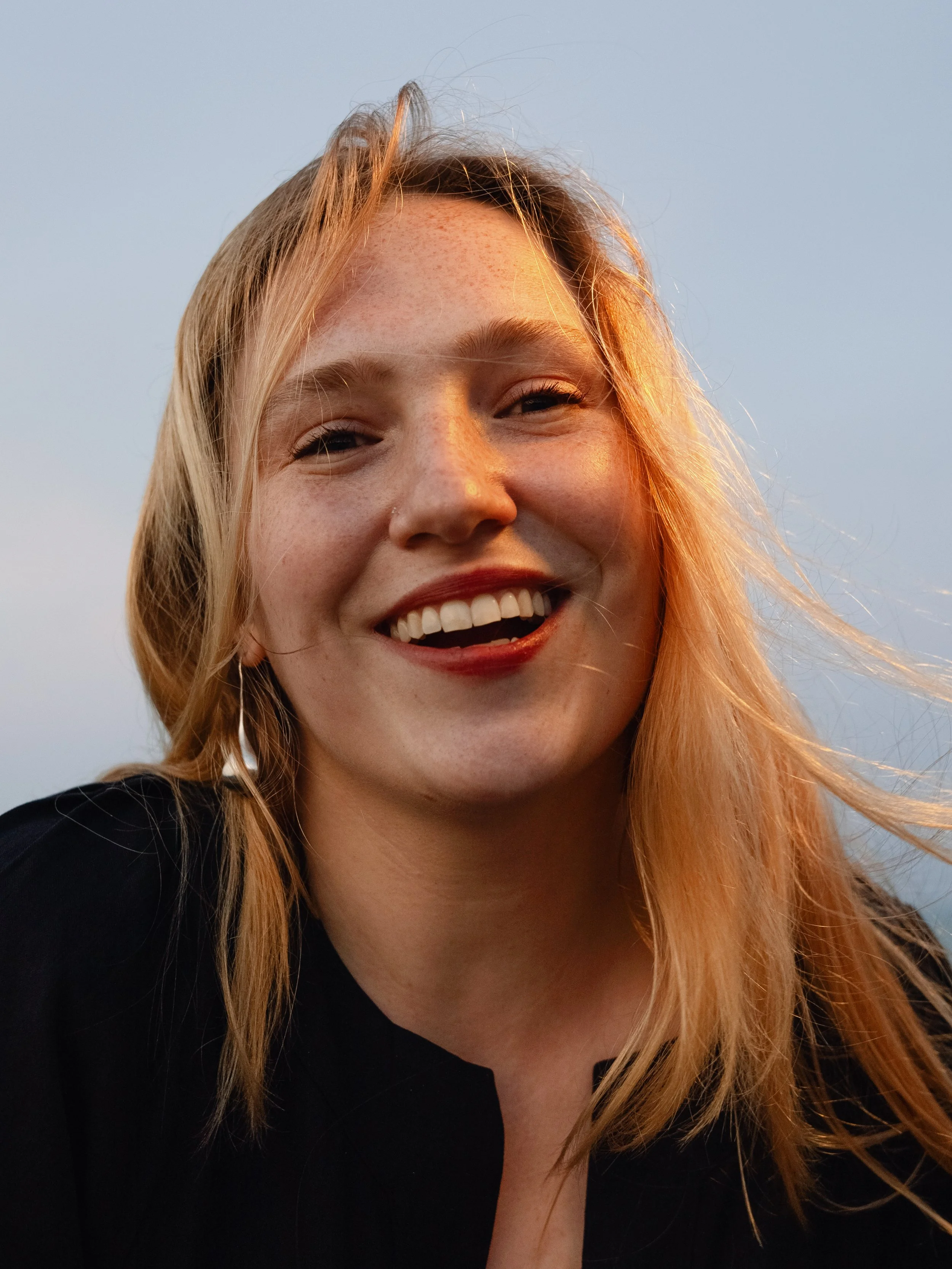 Close-up of a smiling woman with blonde hair outdoors at sunset, wearing a black top.