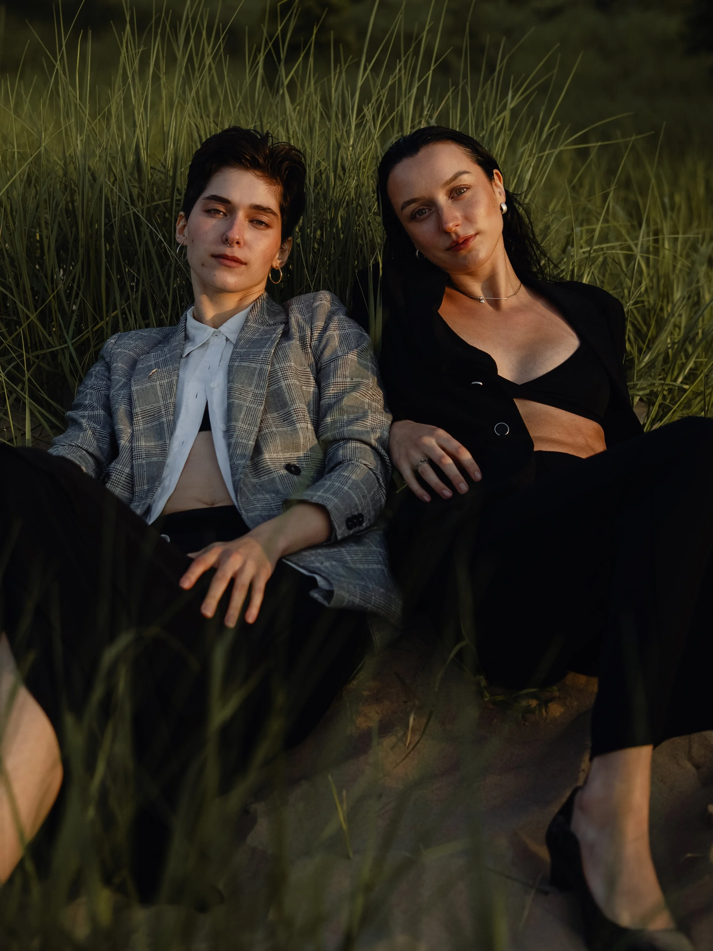 Two women with dark hair lying in tall grass during sunset, one with short hair wearing a plaid blazer and white shirt, the other with longer hair wearing a black top and earrings, both looking at the camera.