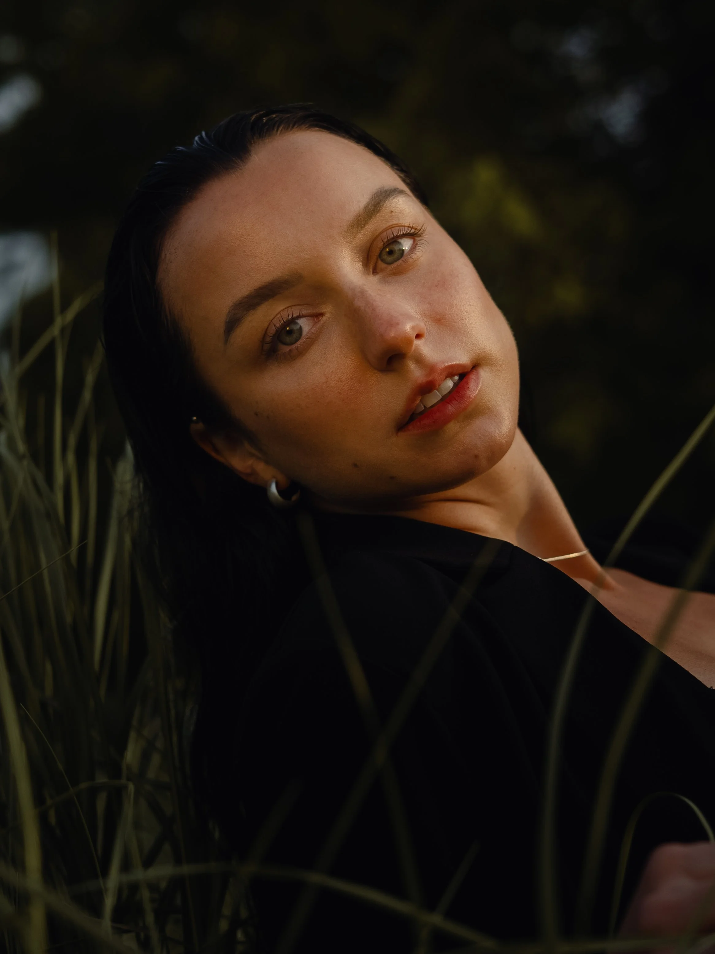 Close-up of a woman's face with her head tilted back, dark hair, and natural makeup, surrounded by tall grass or plants in a dark outdoor setting.