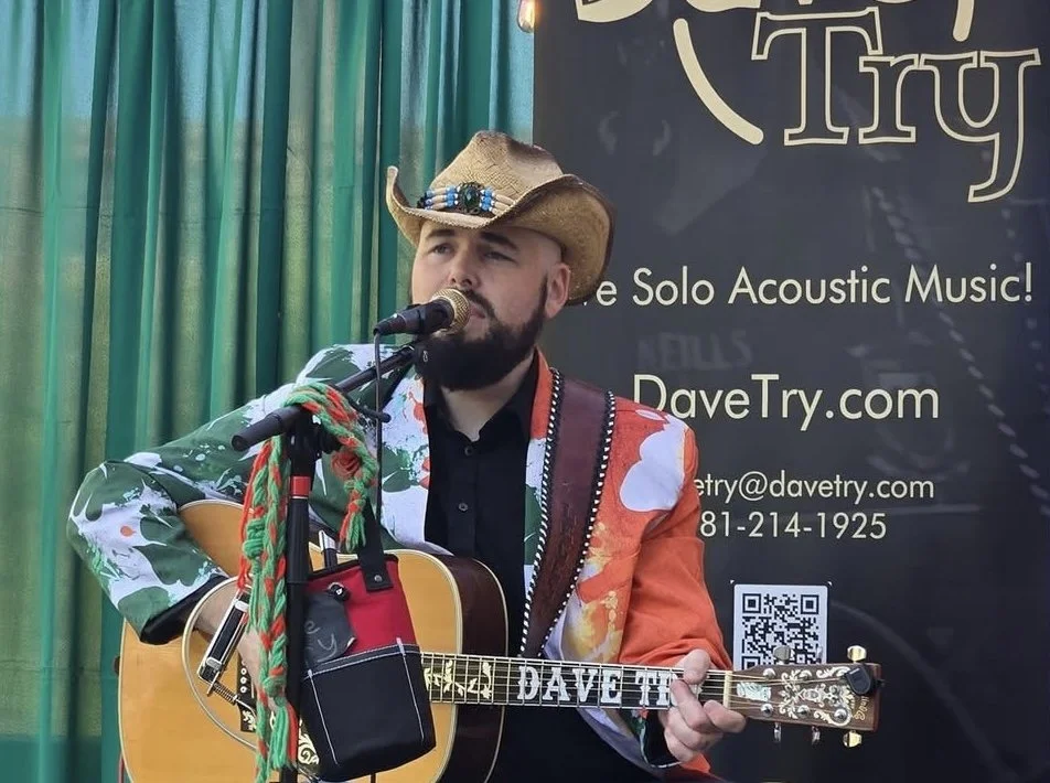 Dave Try performing solo acoustic music, wearing a cowboy hat, Irish jacket, and playing an acoustic guitar in front of a promotional backdrop at an Irish festival.