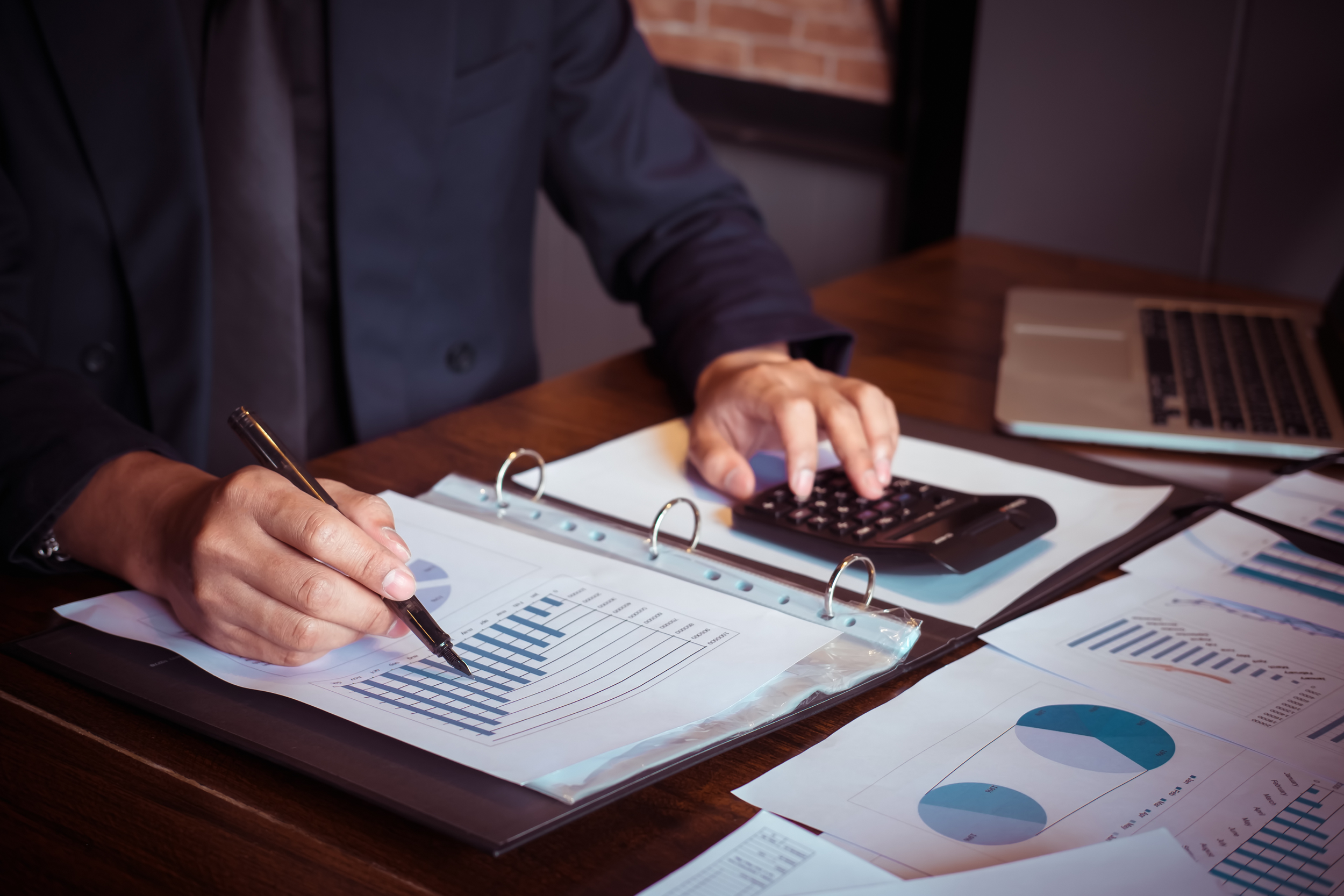 Person reviewing financial charts and graphs with a pen, using a calculator and a laptop on a wooden desk.