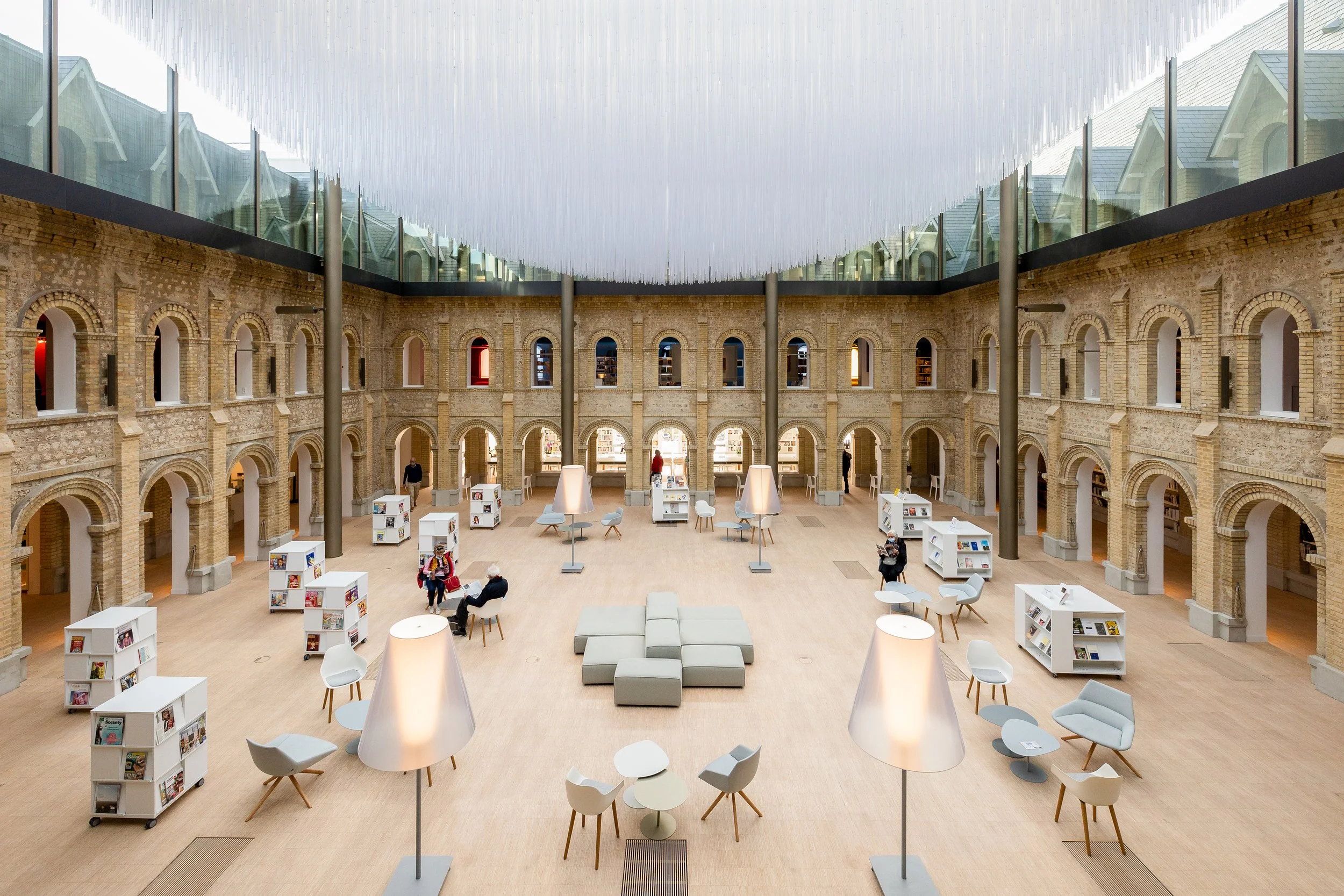 Intérieur moderne d'une bibliothèque ou centre culturel avec arches en briques, mobilier blanc et lampes sur pied, et un plafond de verre au-dessus.