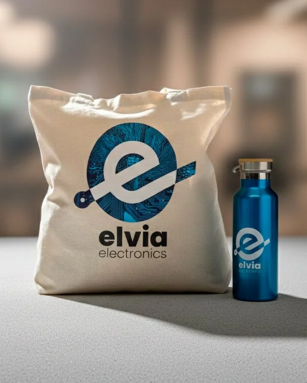 Un sac en toile blanche avec le logo Elvia Electronics et une bouteille en métal bleue avec le même logo, positionnés sur une surface près d'une fenêtre avec un fond flou d'un bâtiment urbain.