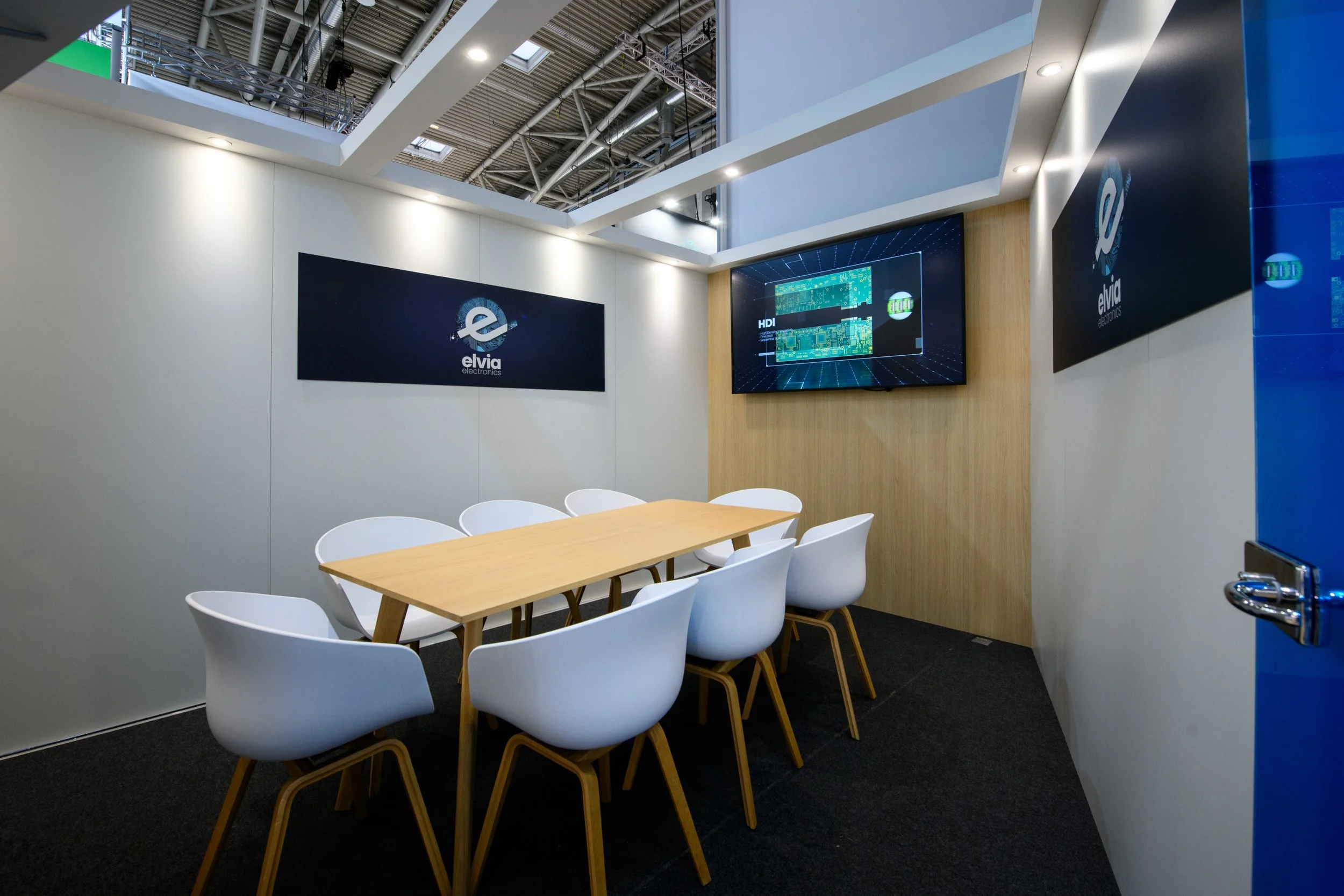 Salle de réunion avec une table en bois clair, six fauteuils blancs, deux écrans vidéos affichant le logo d'Elvia Electronics, plafond en métal apparent, murs blancs avec un panneau en bois clair, sol recouvert de tapis noir.