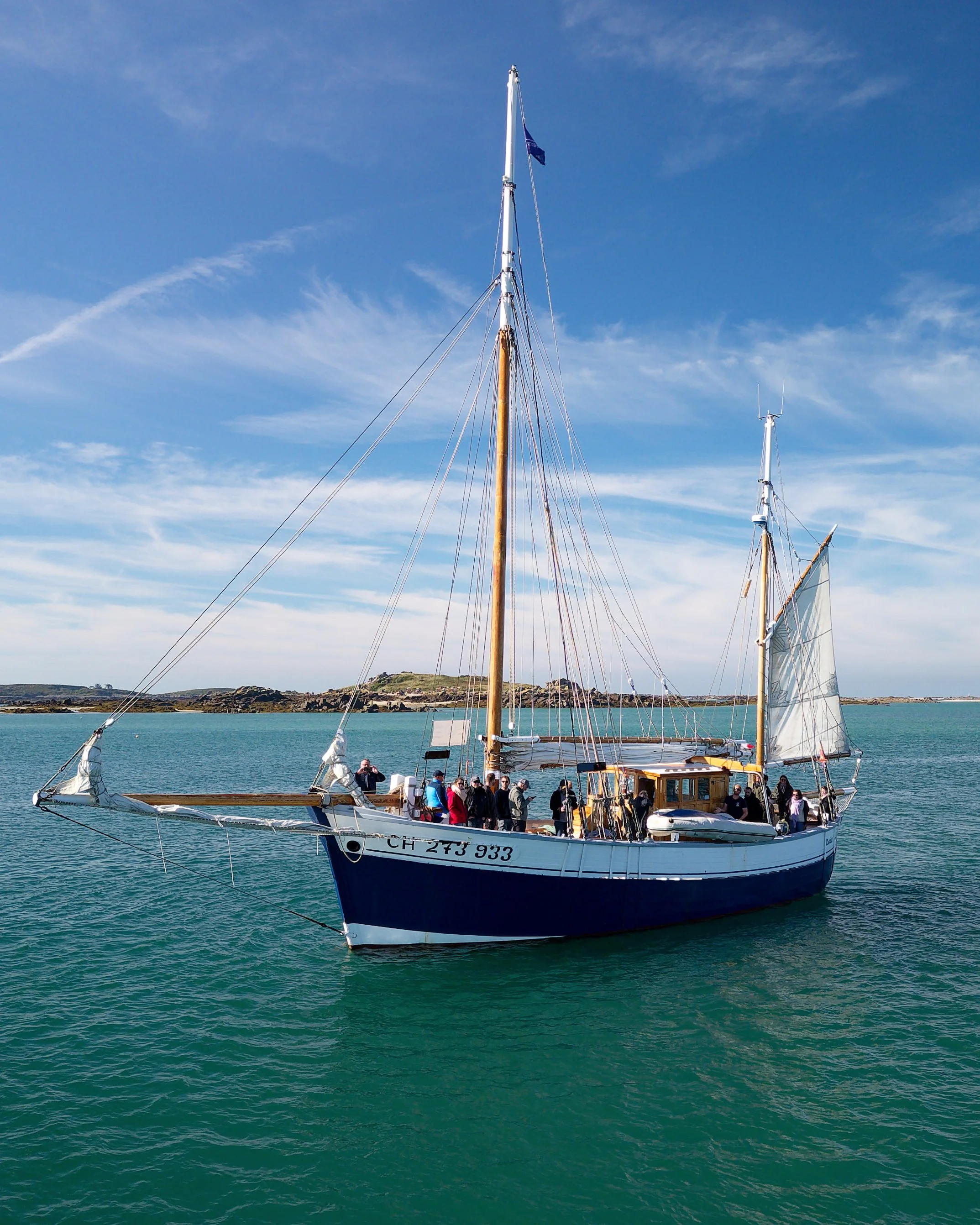 Un voilier avec plusieurs personnes à bord naviguant en mer par une journée ensoleillée, avec un ciel bleu et quelques nuages.