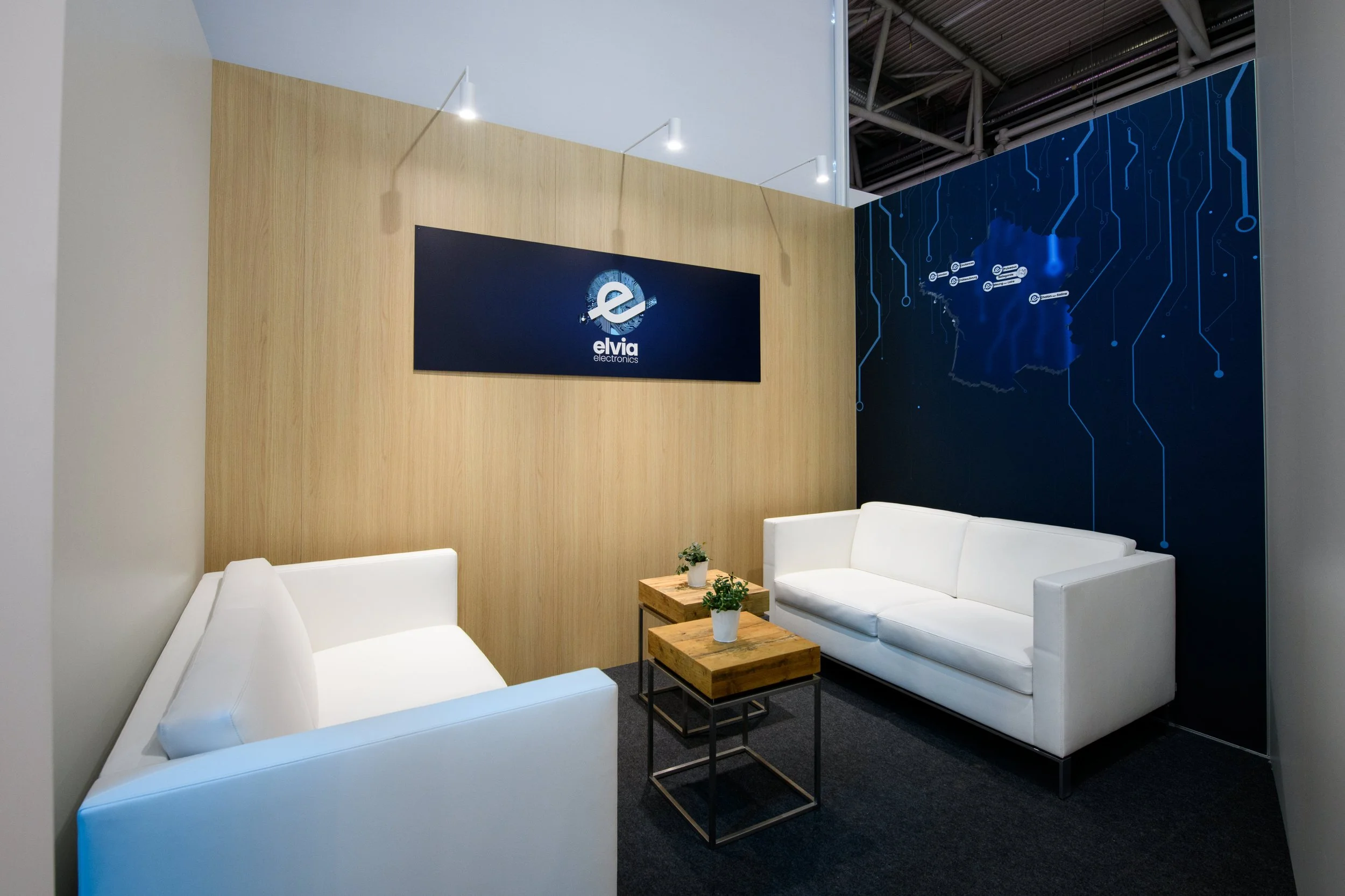 Salle d'attente avec deux canapés blancs, deux tables en bois avec des plantes en pot, écran affichant le logo 'elvia electronics', mur en bois clair et mur en panneau noir avec graphique de carte.'