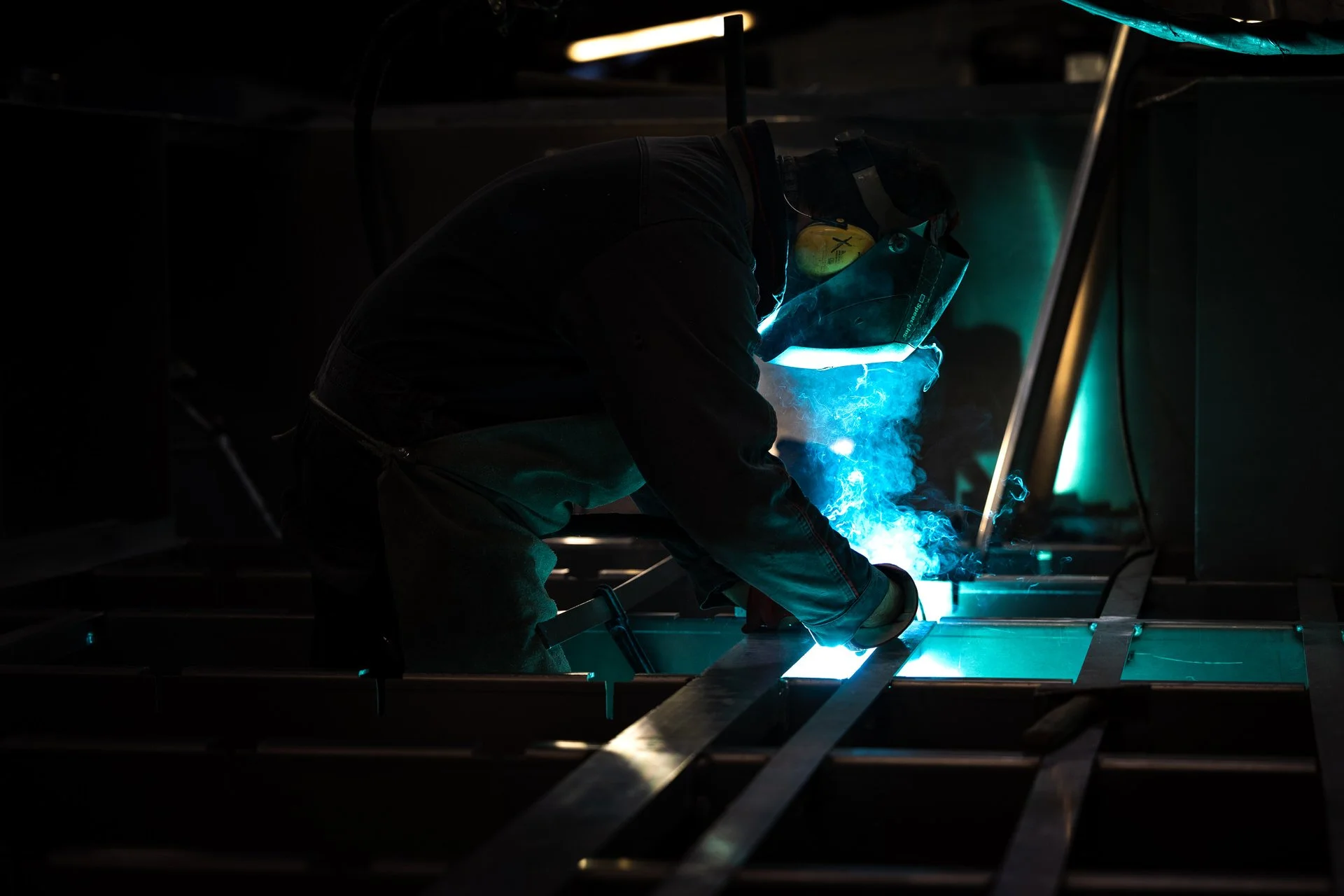 Un soudeur porte un casque avec un masque de protection, travaillant sur une structure métallique dans un environnement sombre, avec des étincelles et une lumière bleue.