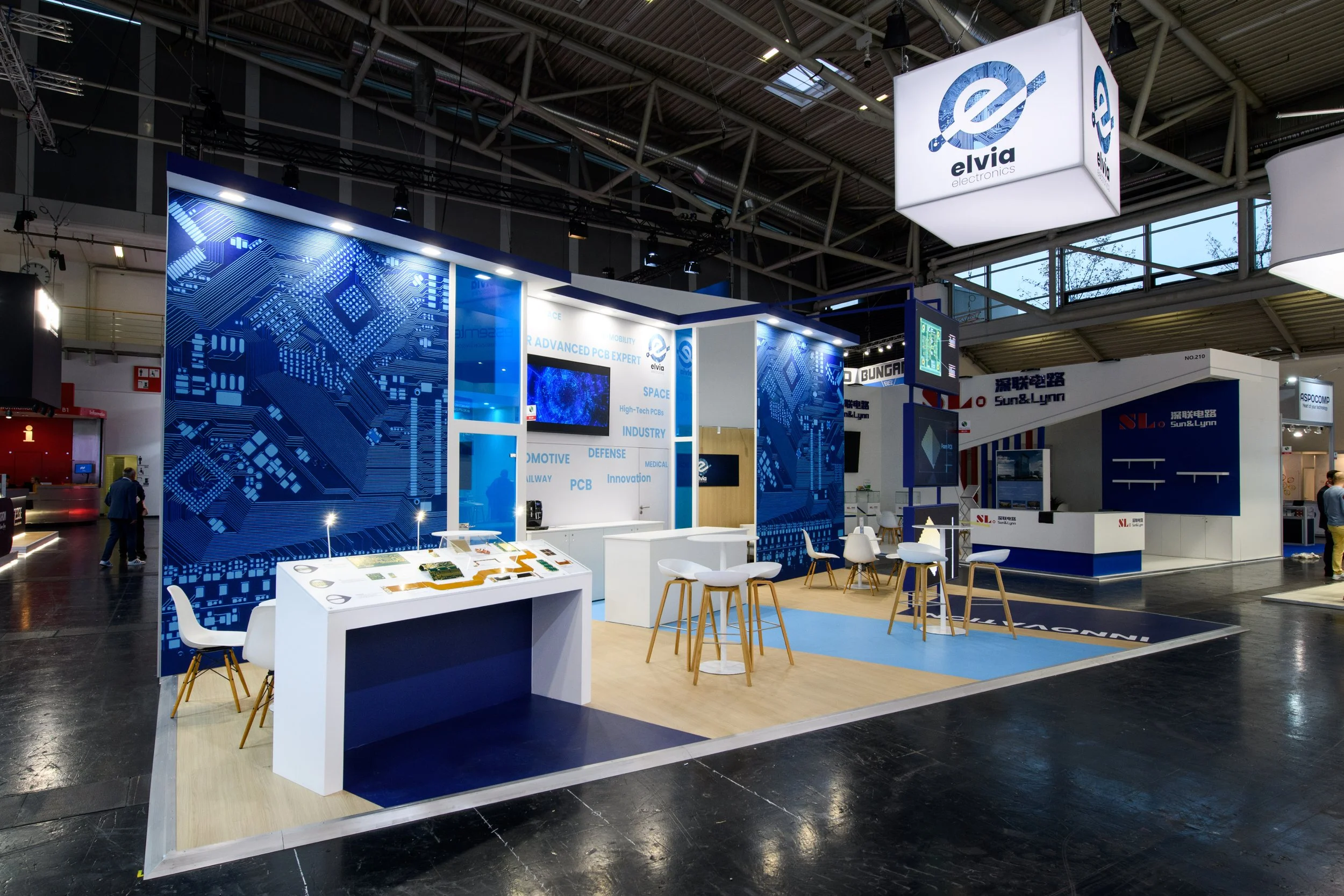 Stand d'une entreprise d'électronique appelée Elvia, présentant des circuits imprimés avancés, avec des tables et des chaises pour des rencontres, dans un salon ou une foire d'exposition.