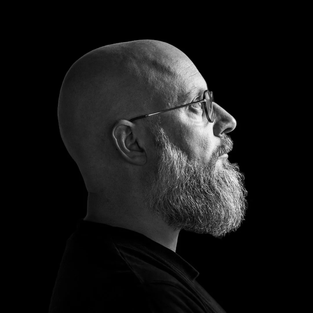 Portrait en noir et blanc d'un homme chauve, avec une barbe grise, portant des lunettes, vu de profil contre un fond noir.