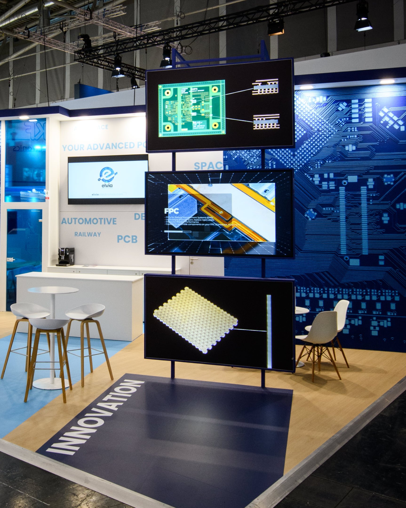 Un stand d'exposition mettant en avant des composants électroniques, avec trois écrans affichant des circuits imprimés et des éléments électroniques, dans un environnement technologique et moderne.