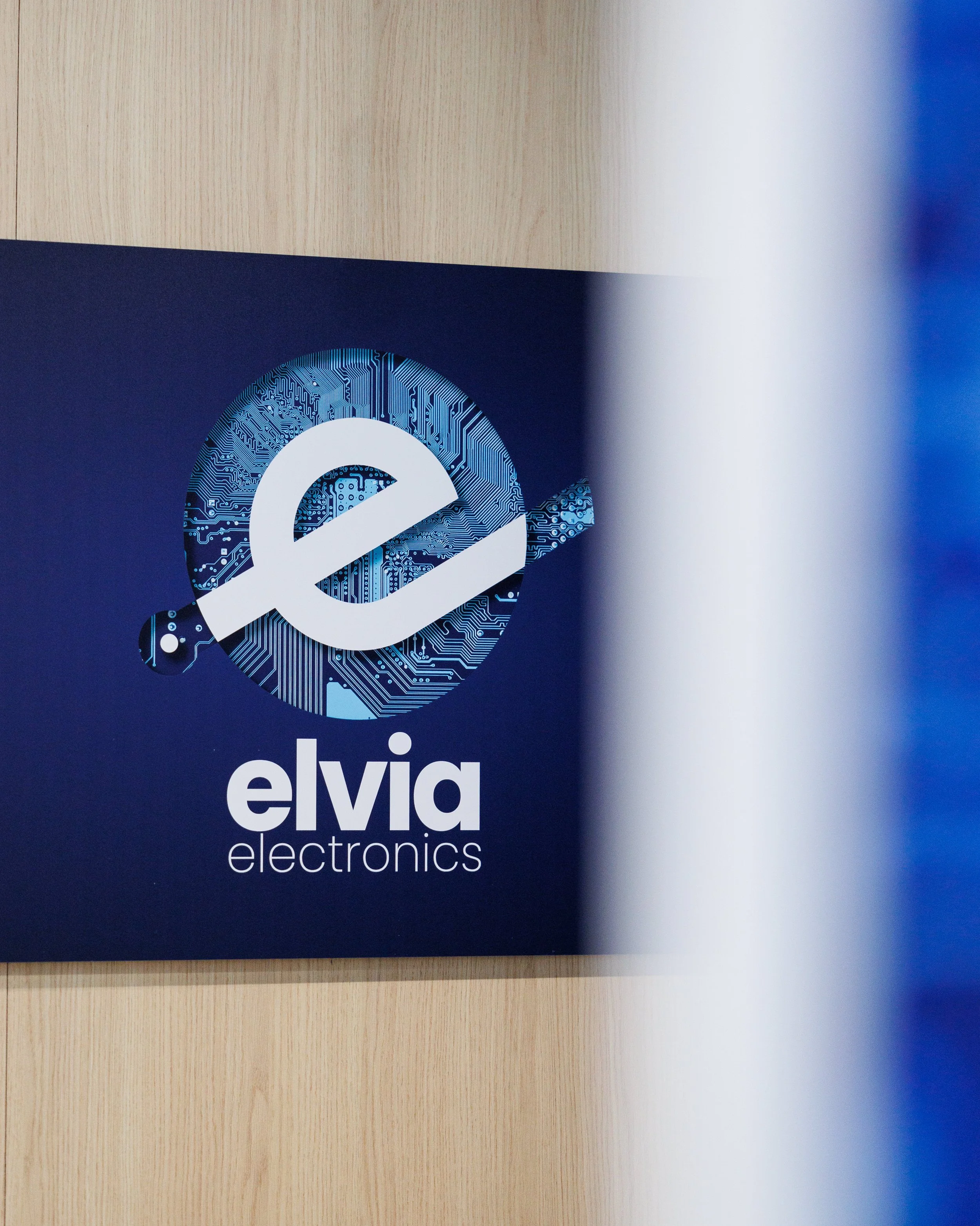 Logo d'une entreprise appelée Elvia Electronics avec un symbole représentant une électrode ou un composant électronique sur un fond bleu avec des motifs de circuits imprimés.