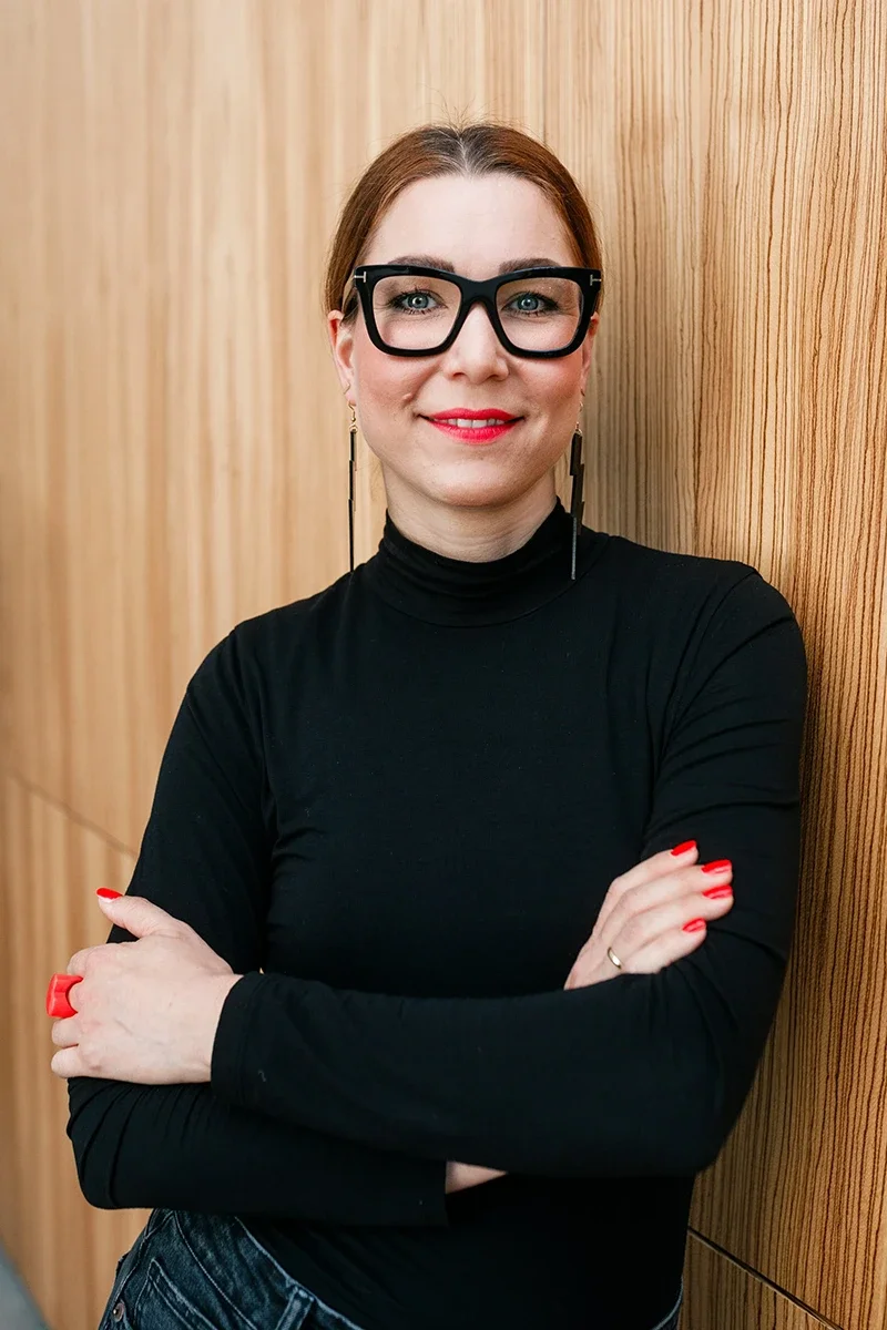 Frau mit kurz braunen Haaren, schwarzen Brillen, rotem Lippenstift und roter Nagellack, trägt schwarzes Turtleneck-Shirt, steht vor einer Holzvertäfelung, lächelt.