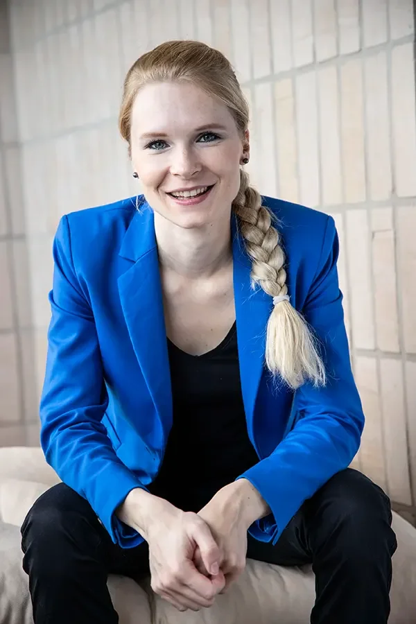 Marie Schlierkamp mit blonden Haaren, in einem blauen Blazer, sitzt vor einer Steinmauer und lächelt in die Kamera.