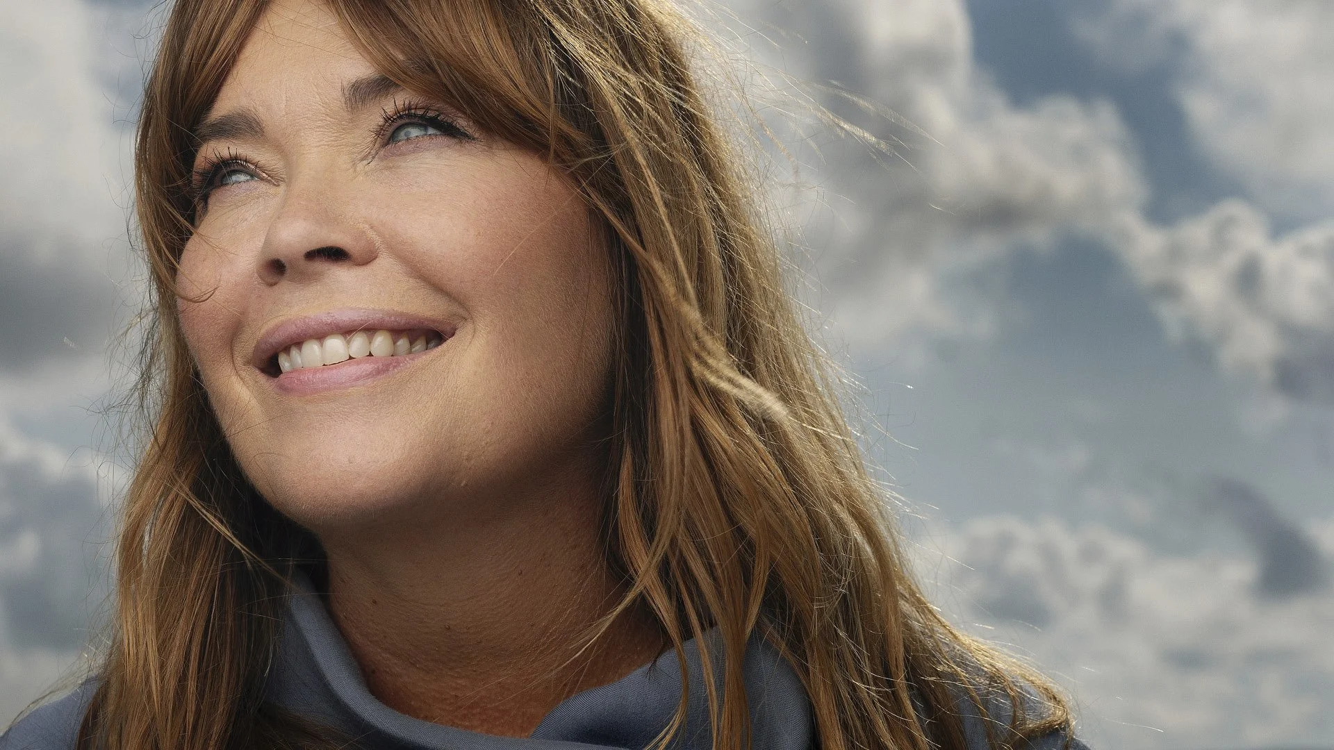 Solveig Slettahjell smiler og ser bort med glimt i øyet, med skybelagt himmel i bakgrunn.