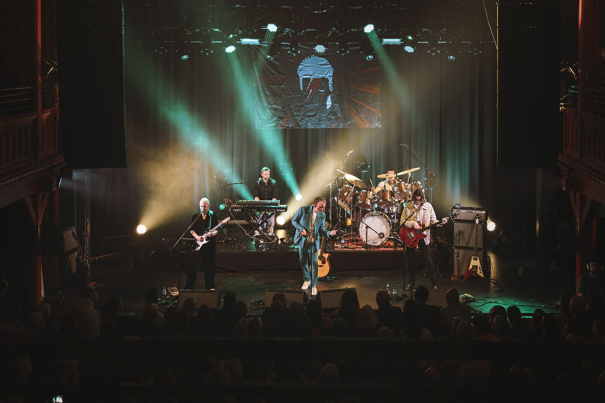 The Spiders From Mars hyller David Bowie i et fullsatt Byscenen. En stor David Bowie illustrasjon henger på veggen bak bandet.