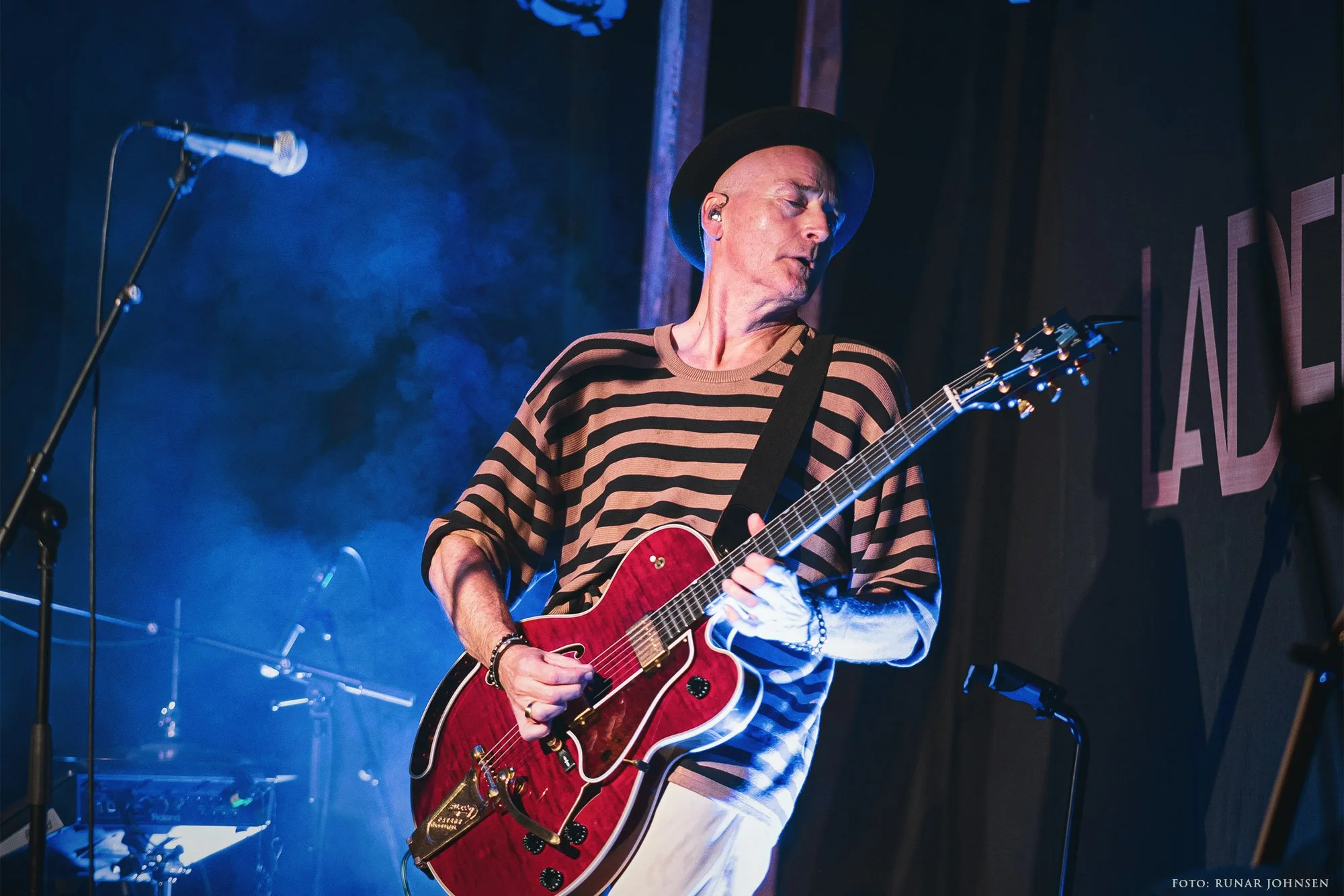 Morten Abel med rød gitar på Ladekaia i Trondheim, omgitt av blått scenelys og røyk