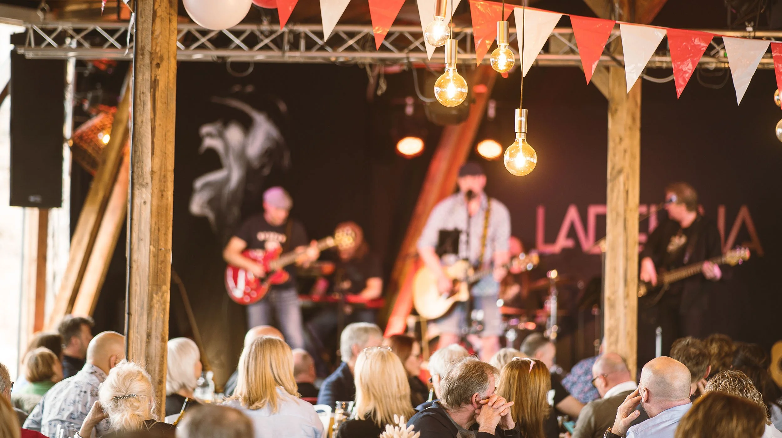Publikum ser på en live musikkonsert på en innendørs scene med tre musikere som spiller gitar. Dekorative lamper og flagg i rødt og hvitt henger over scenen.