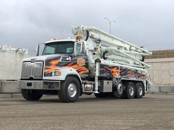 Concrete pump truck with a white cab, black and orange flame-like graphics, and large white concrete pumping pipes on a paved surface.