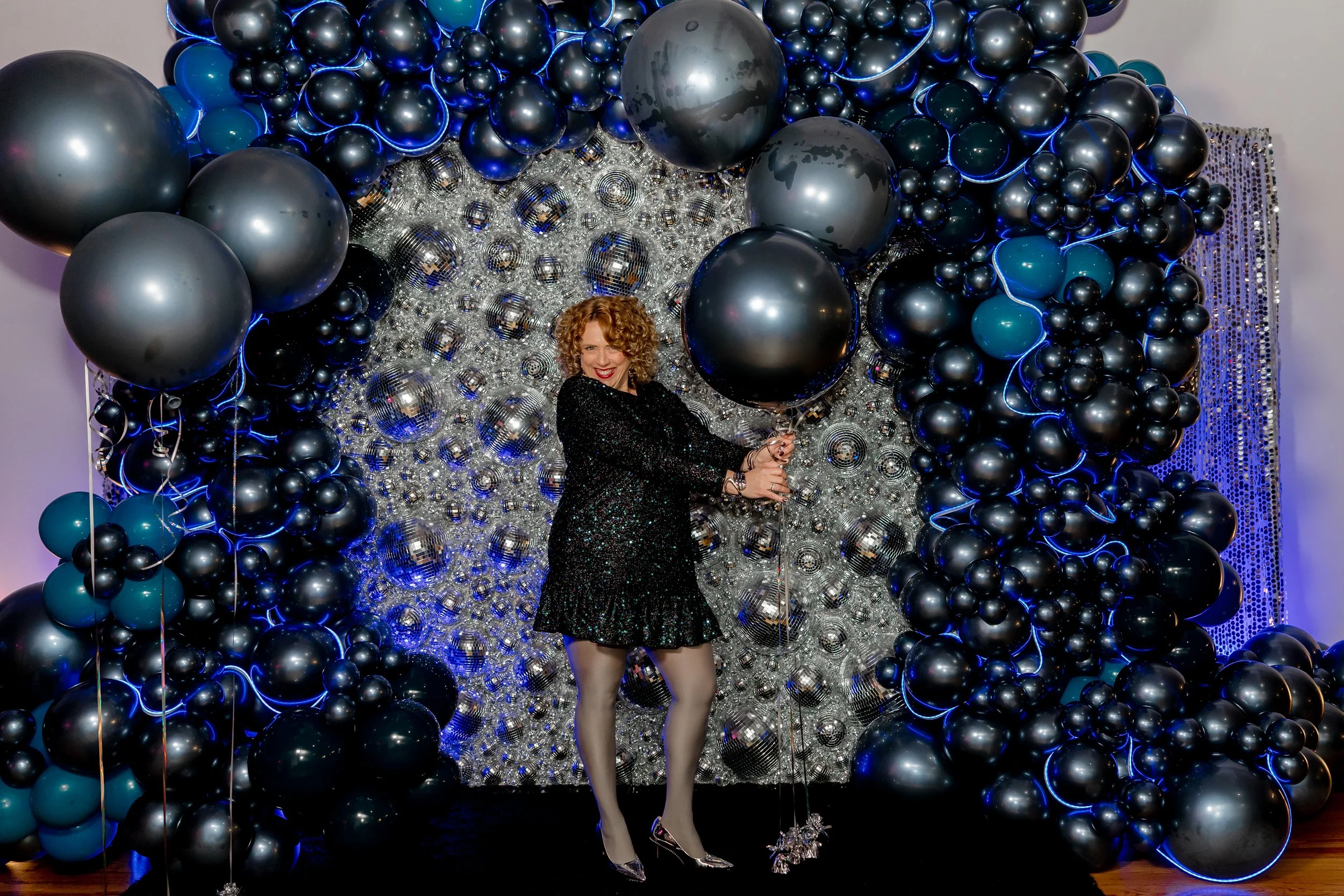 Woman posing in front of a large black, silver, and blue balloon installation featuring metallic orb balloons and a disco-ball-style reflective backdrop, holding oversized dark balloons and smiling in a glamorous event-style balloon display.