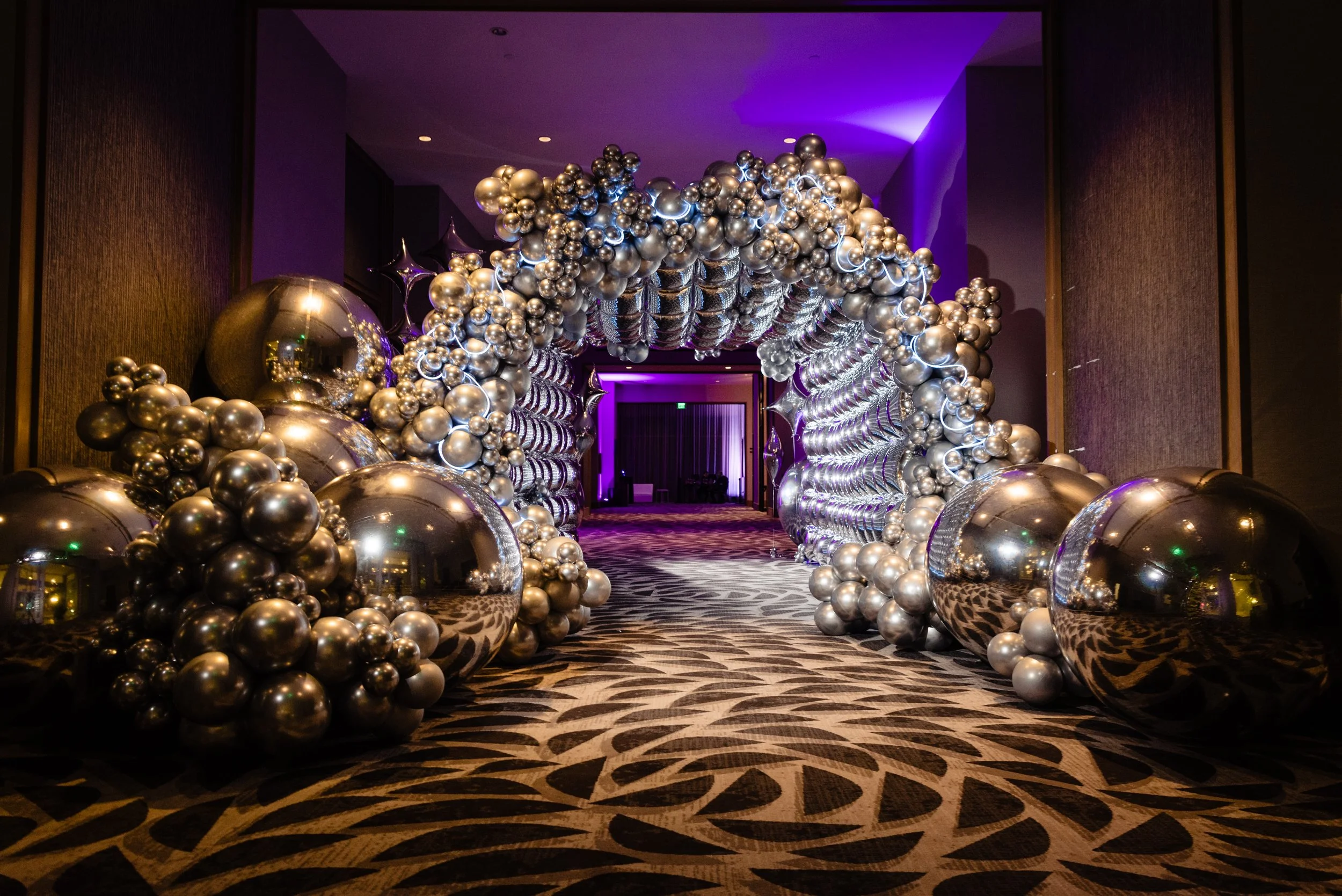 An ornate balloon arch made of silver and reflective square metallic balloons, decorated with star-shaped balloons, creating an elegant entryway at Houston Space Center's Holiday Party