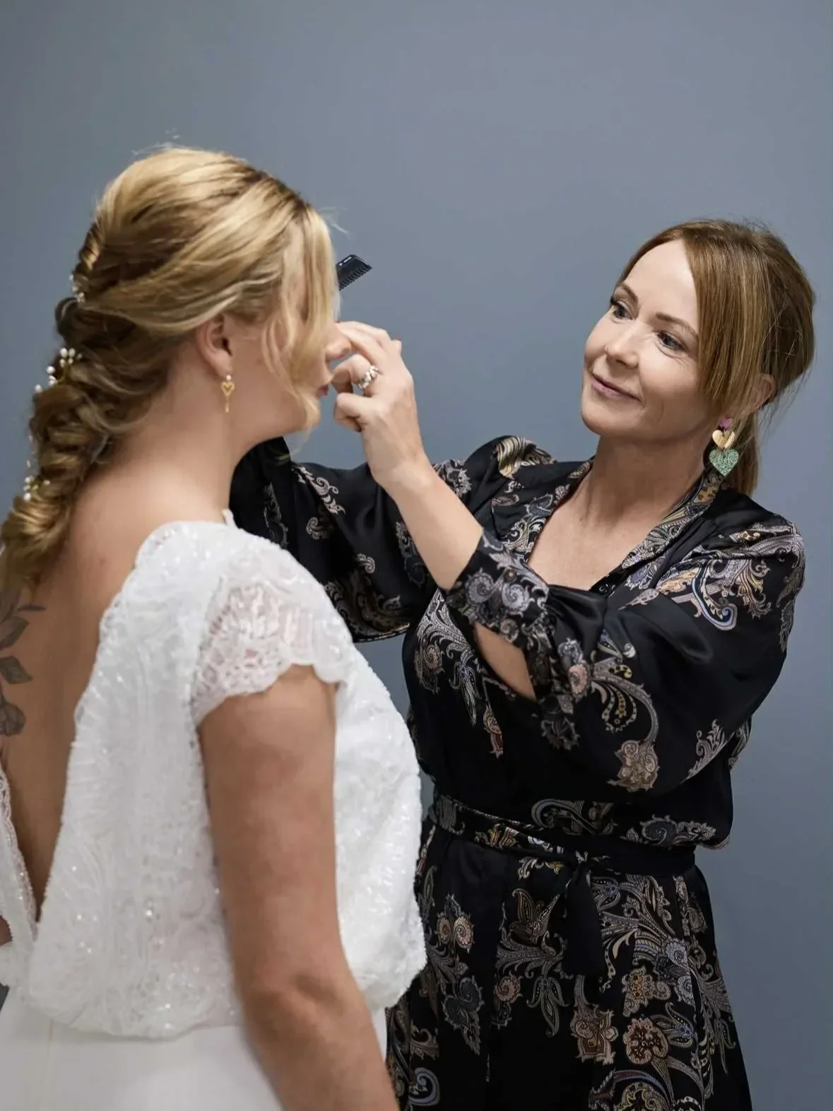 Frisør står med ansigtet til kamera imens hun retter de sidste detaljer på brudens hor og makeup. Bruden står i brudekjole med ryggen til og smuk fletning og perler i håret