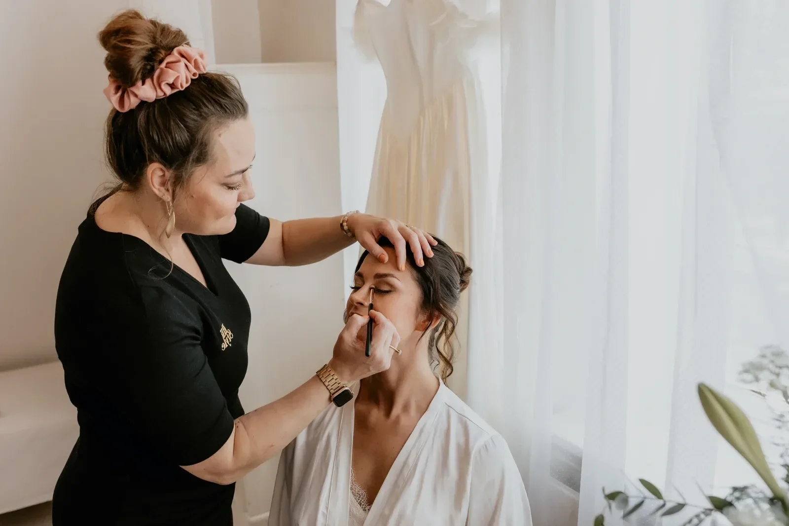 makeupartist i sort kjole og med opsat hår retter øjenskyggen på bruden som sidder foran sin brudekjole