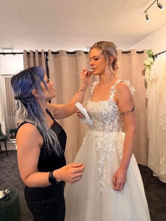 brudestylist og makeupartist som sort tøj og blåt hår giver bruden i brudekjole læbestift på