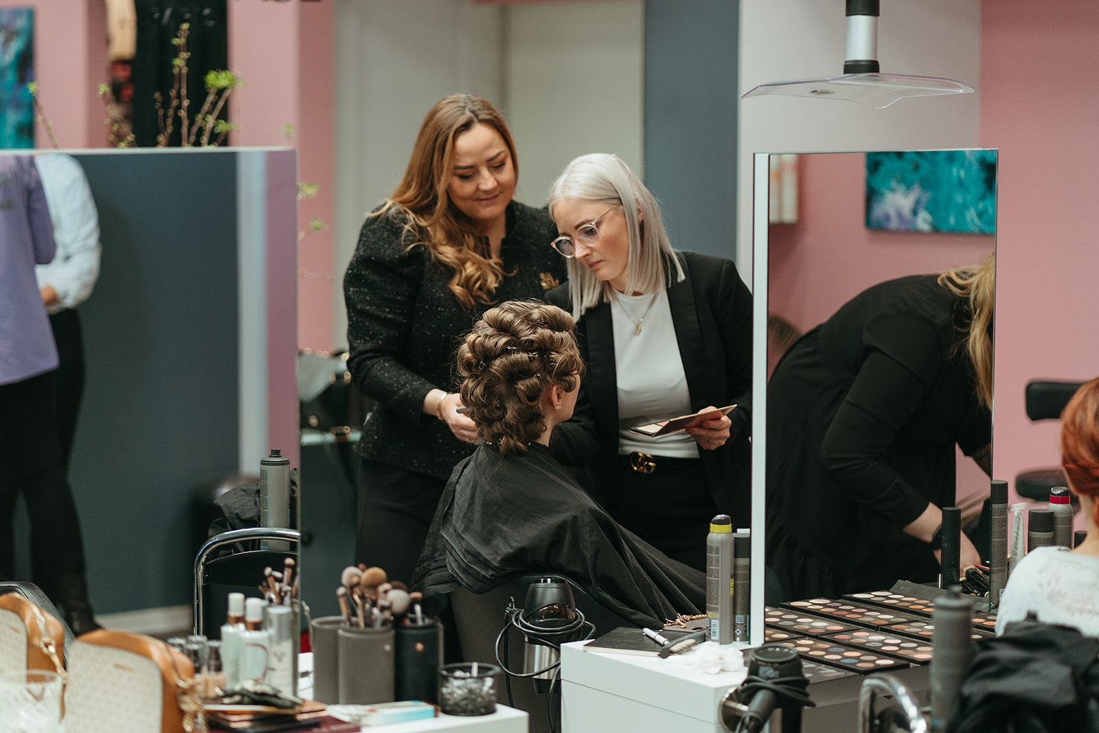 Kvinde med rollet hår bliver forberedt i en makeup-station, mens to kvinder taler med hende. Der er forskellige makeupprodukter og værktøj på bordet.