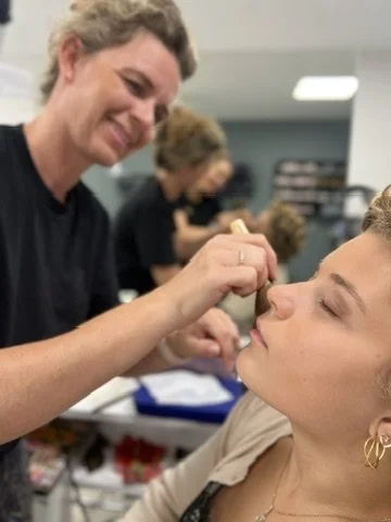 Smilende frisør lægger makeup på naturlig ung skandinavisk kvinde med guldøreringe