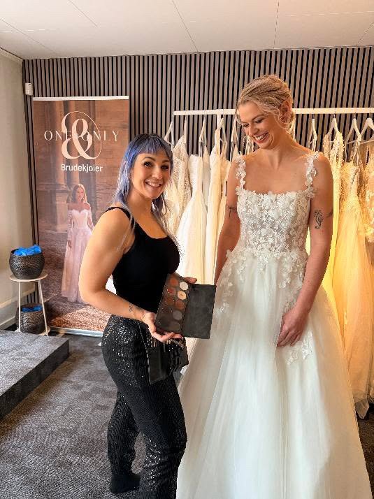 frisør og brud står ved siden af hinanden i brudekjolebutik. Stylisten holder en makeuppallette og begge kvinder smiler