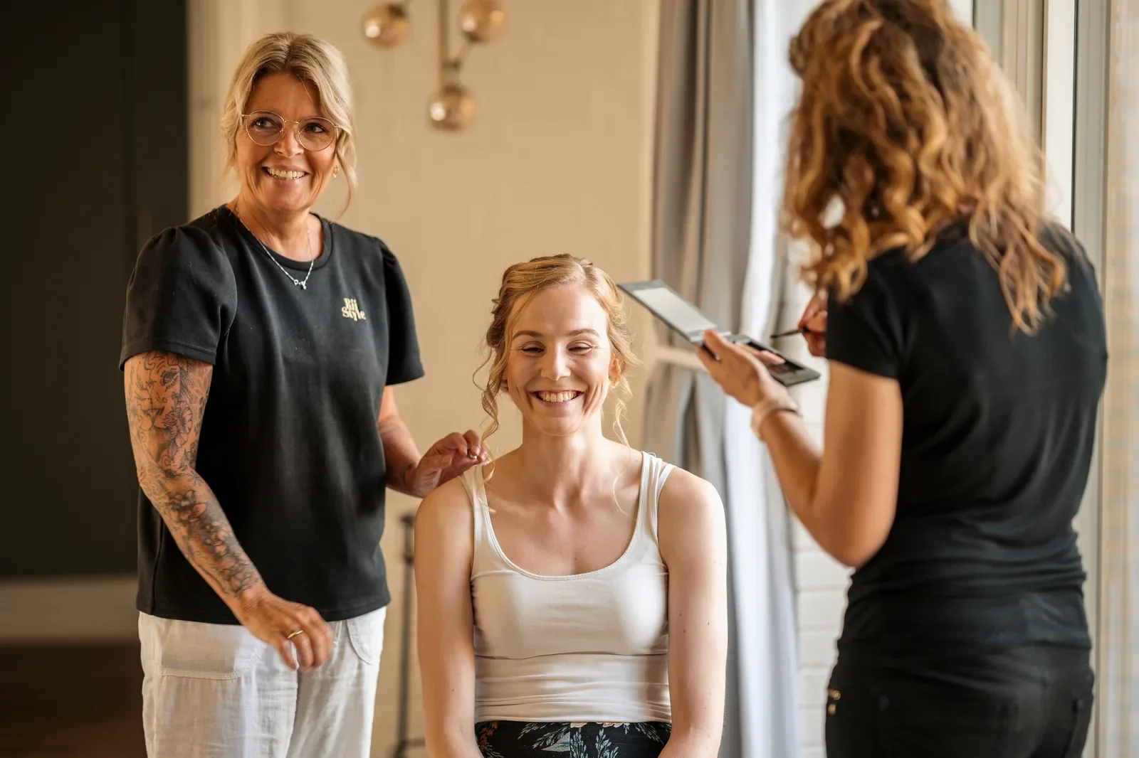 To stylister er igang med forberedelserne af hår og makeup på bruden. Den ene stylist står med en rolig hånd på brudens skulder og den anden stylist med makeuppalletten klar til brudemakeuppen